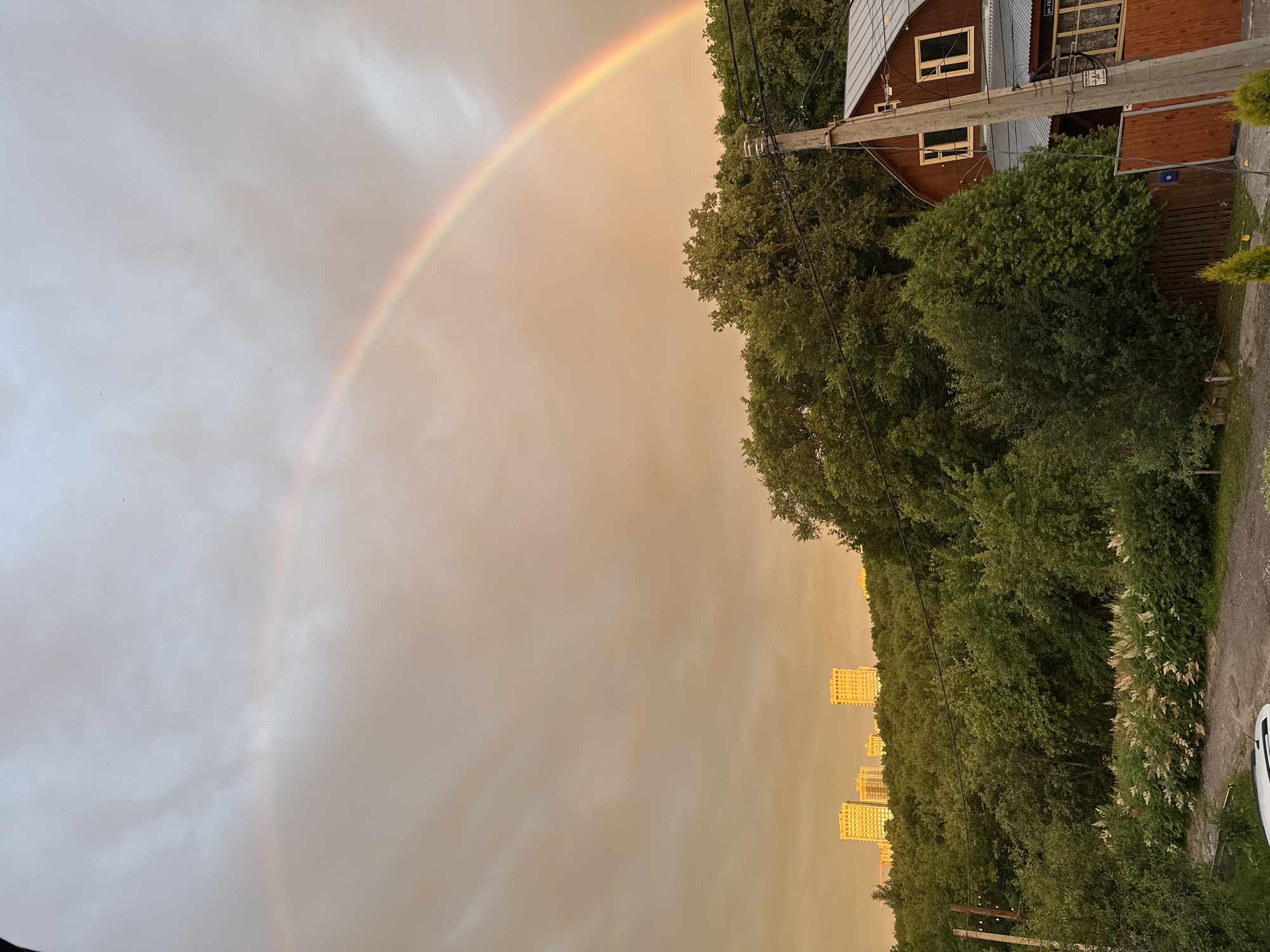 The image depicts a panoramic view of a suburban landscape on a cloudy day, featuring a prominent double rainbow arching across the sky. In the background, several modern, light-colored high-rise buildings are visible, suggesting an urbanized area. These buildings are situated atop a densely forested hill. In the foreground, a rustic wooden house with a corrugated metal roof and visible windows stands amidst lush greenery and trees. A wooden fence and a utility pole with electrical wires are also present near the house. A dirt path winds through the scene, with a white car parked partially visible at the bottom left. The sky is overcast with a warm, diffused light, indicative of either sunrise or sunset, which contributes to the soft colors of the rainbow. This scene is consistent with the suburban outskirts of Nizhny Novgorod, Russia, where modern high-rise developments often coexist with more traditional residential areas and natural landscapes.