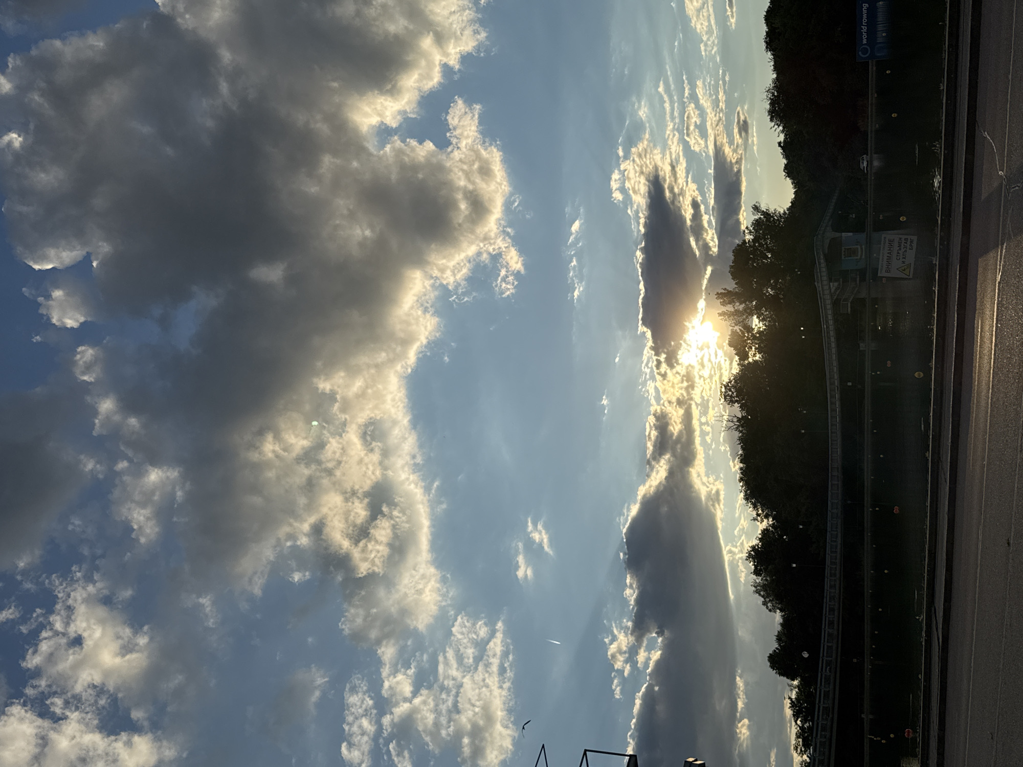 The image depicts a sunset over a body of water, likely a river or canal, bordered by trees and a bridge. The sky is filled with dramatic clouds, illuminated by the setting sun, which creates rays of light piercing through the clouds. In the foreground, a road is visible. Along the waterfront, there is a low structure with some warning signs and a logo that appears to be for "world rowing". The scene suggests a tranquil, natural setting within or near a city. Given the "world rowing" signage, the location is likely a rowing canal or a water body used for rowing. Plovdiv, Bulgaria, is known for its rowing facilities, particularly the Plovdiv Rowing Canal, which hosted numerous international competitions. This image is consistent with a view of that location during sunset.