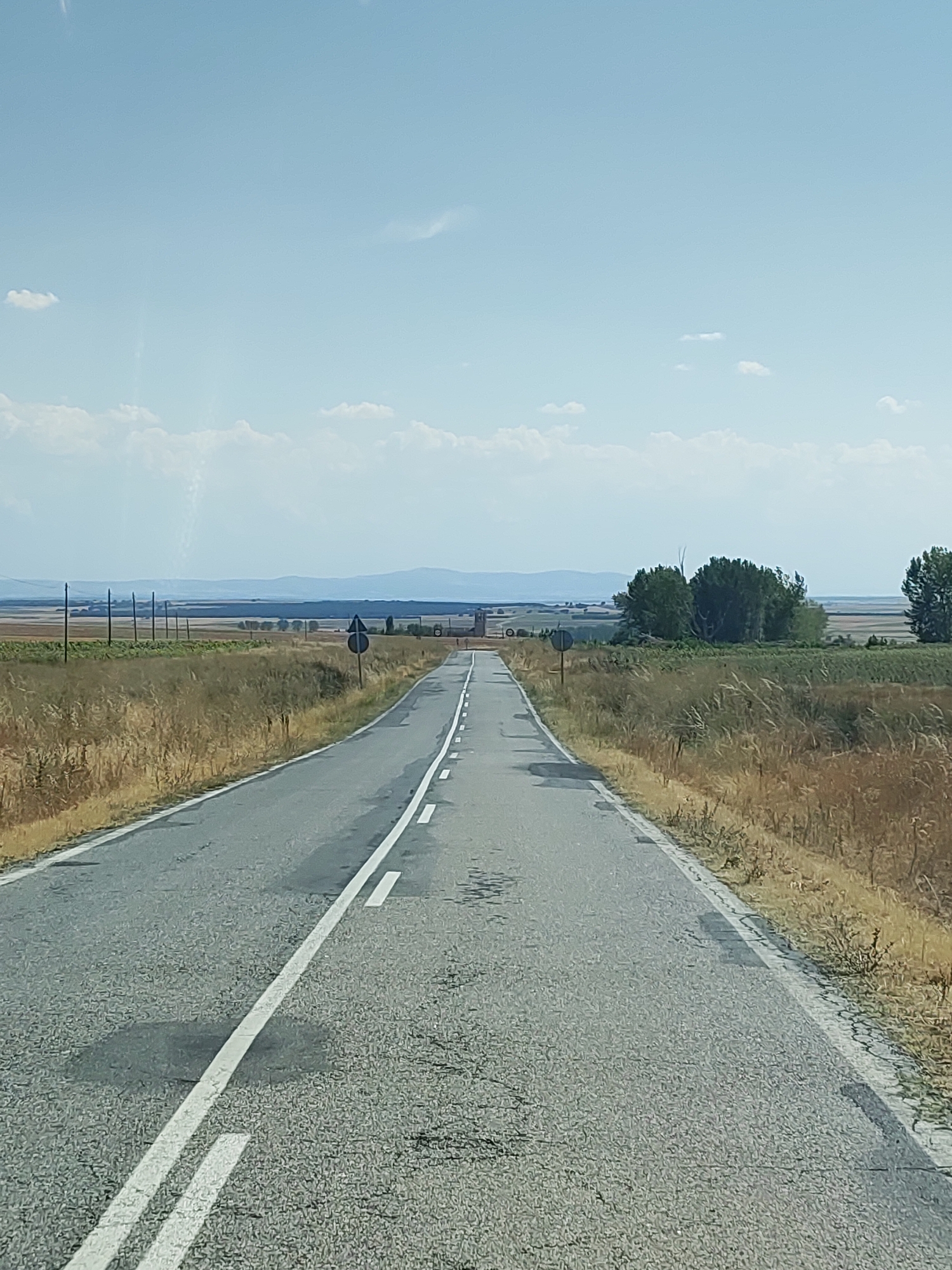 The image shows a straight asphalt road in a rural area, likely in Spain, given the context of Santa María la Real de Nieva. The road is divided by a white double line. On either side of the road, there are dry, grassy fields. In the distance, there are scattered trees and what appears to be a long range of hills or mountains under a bright blue sky with some scattered white clouds. There are no people or vehicles visible in the image. The scene suggests a warm, dry climate.