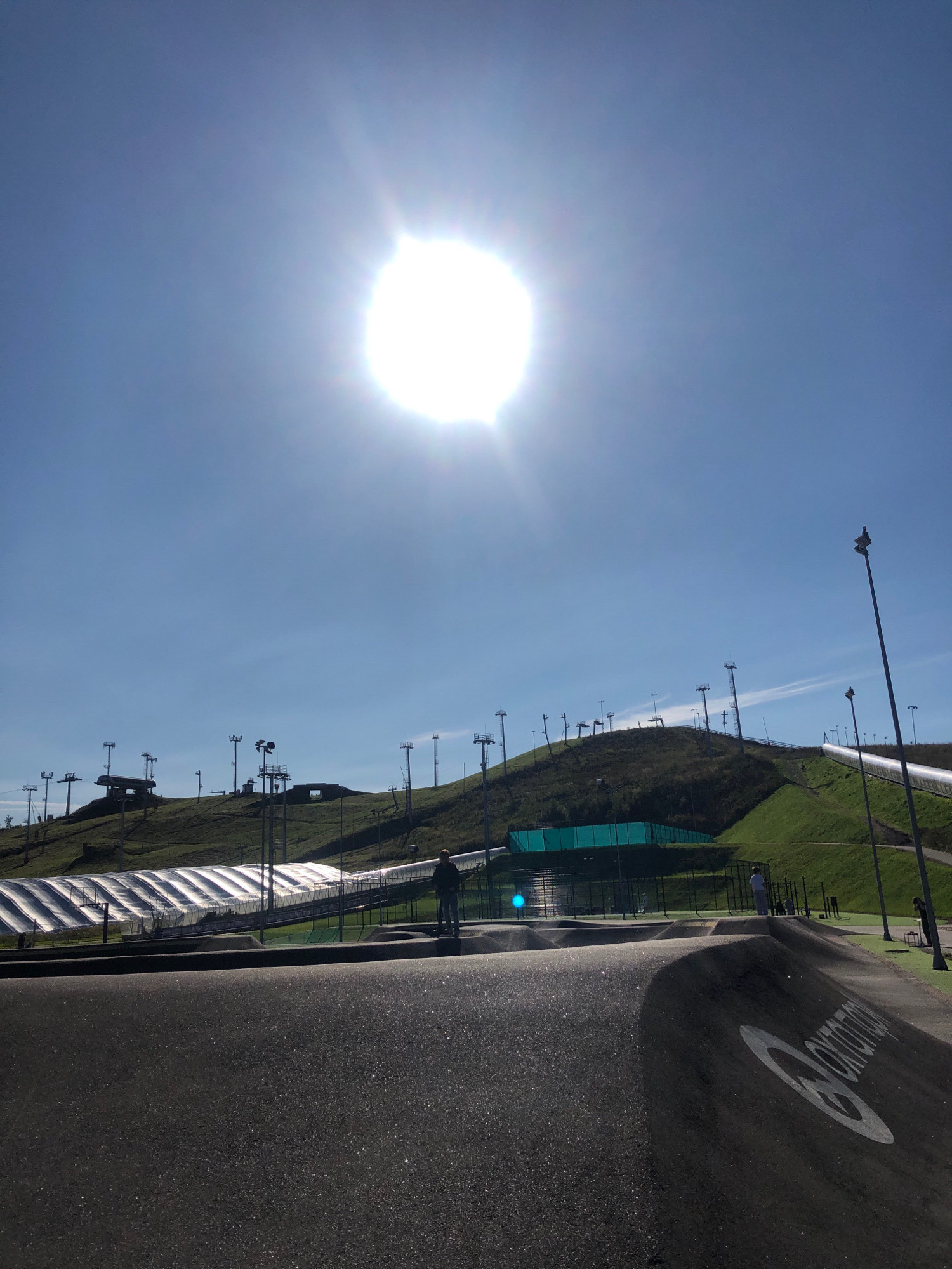 In Okhtinskiy Park, Russia, a bright sun shines from the upper left quadrant of the sky, casting rays over a green, grassy hillside. The hillside is dotted with numerous tall, slender light poles. At the top of the hill, a ski lift structure is visible. Near the base of the hill, a long, translucent, tent-like structure stretches horizontally. In the foreground, a black asphalt pump track is depicted, with a white circular logo visible on its surface. Two individuals are present: one man stands on the right side of the pump track, and another person is seen on the grassy area near the translucent structure on the hillside. The sky is a clear, bright blue, with a few faint streaks of clouds.