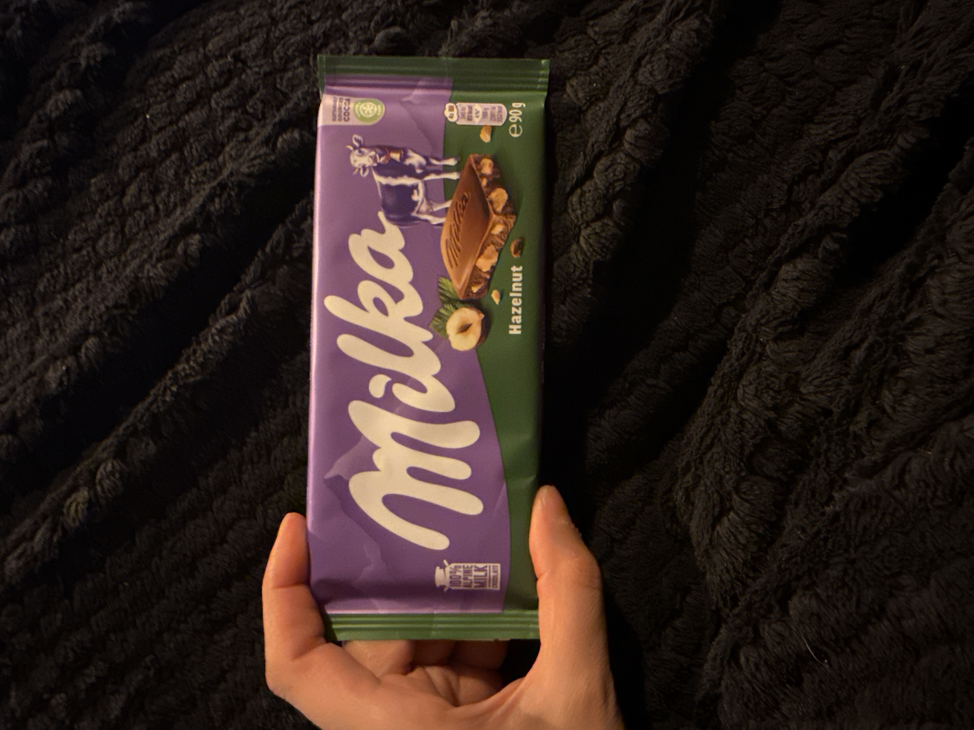 A human hand with light skin tone is visible on the left side of the frame, holding a Milka Hazelnut chocolate bar. The bar's wrapper is predominantly purple with a green lower section, featuring the white "Milka" logo, an image of a cow, and a visual representation of the hazelnut chocolate bar, including loose hazelnuts. Textual elements visible on the wrapper include "100% ALPINE MILK," a symbol for "Sustainably Sourced Cocoa," and "e 90g" indicating net weight. The background is a dark, plush, or fuzzy textile material, likely a blanket or throw, exhibiting folds and variations in texture. This scene is located in Great Yarmouth, United Kingdom.