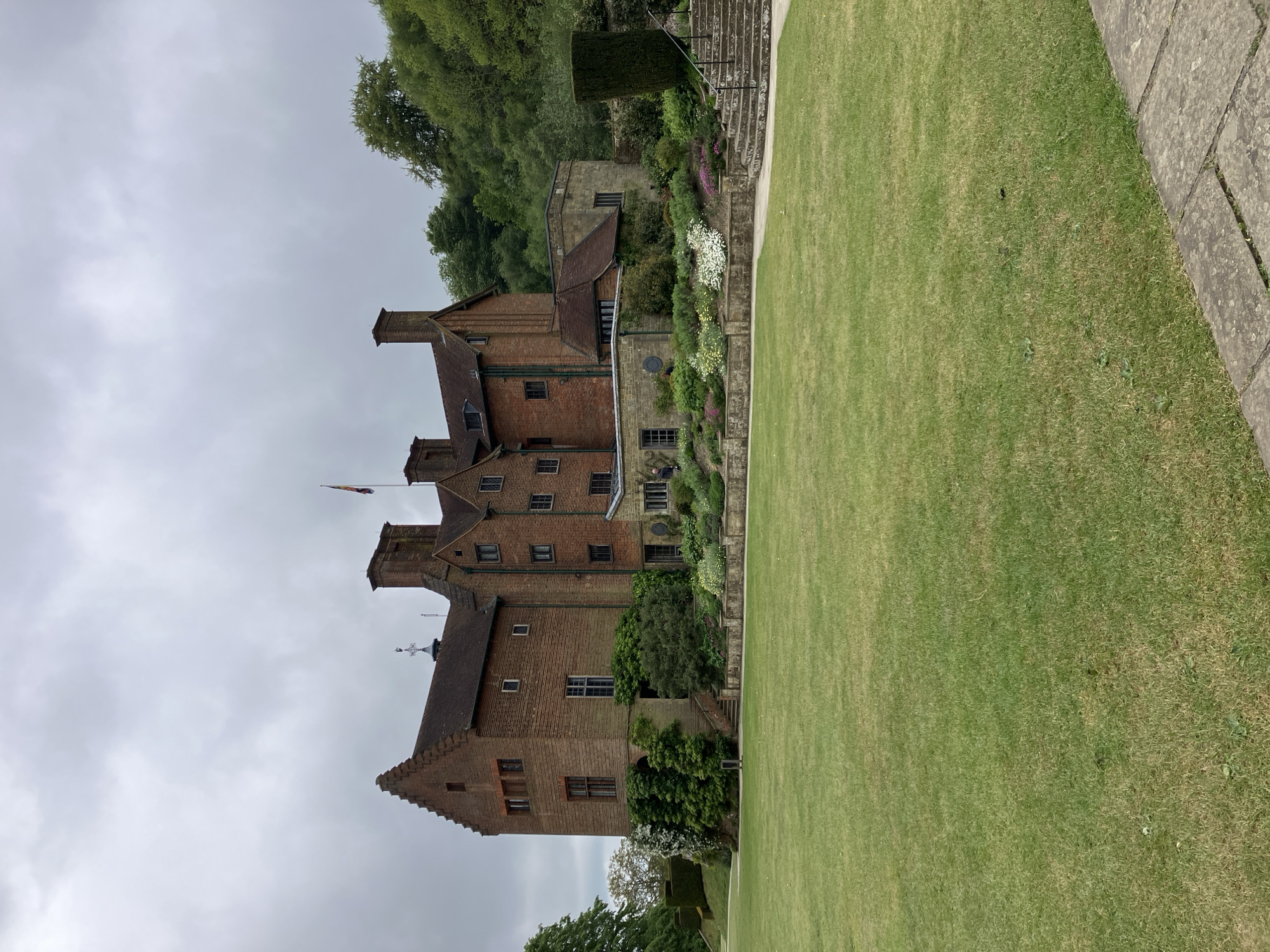 The image shows Knole House, a country house near Sevenoaks, Kent, United Kingdom. The large building is constructed of red brick and has multiple gabled roofs. A flag is visible atop one of the towers. Adjacent to the house is a well-manicured lawn and terraced gardens with flowers and greenery. Stone steps are seen on the side of the building. The sky is overcast.