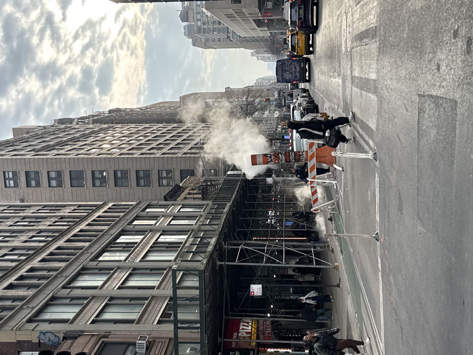 A city street scene features steam rising from a utility vent surrounded by orange and white traffic barriers. Scaffolding covers a building facade, while pedestrians walk below. Traffic is visible in the background, including cars, taxis, and a graffiti-covered van. Buildings line both sides of the street, reaching up towards a cloudy sky.