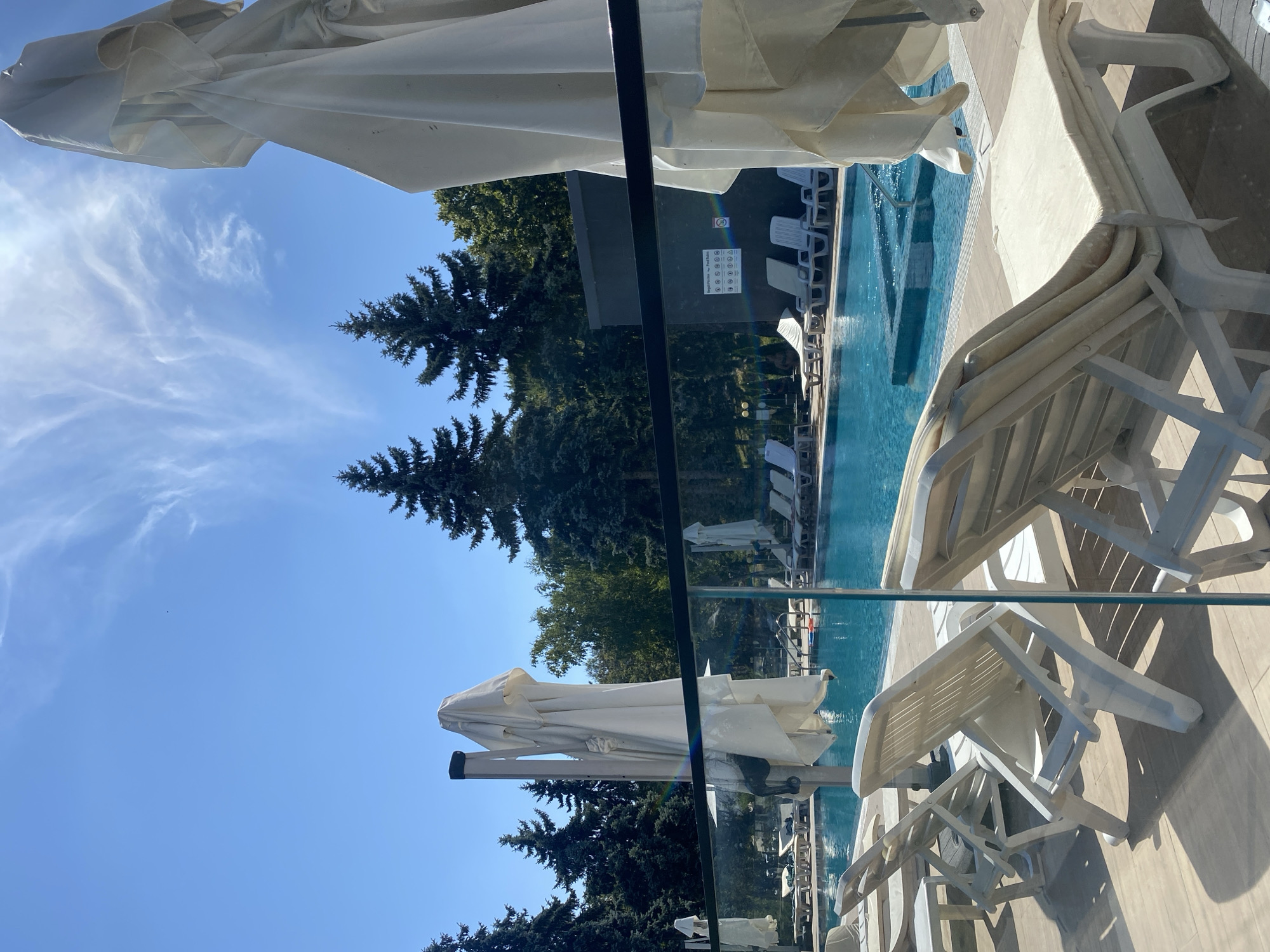 A bright, sunny day is depicted at a swimming pool area, likely in or near the city of Ustia, Moldova, given the context. The foreground is dominated by several white plastic lounge chairs, some folded and stacked, others set up and ready for use, casting long shadows on the wooden deck. A partial view of a shimmering blue swimming pool with steps and a jacuzzi area is visible beyond the chairs.
Several large, white beach umbrellas, currently closed and covered, stand prominently. One is directly in the foreground, to the right, its fabric draped and partially obscuring the view. Another is in the middle ground, to the left, on a tall pole. In the background, a dense line of tall, dark green pine trees forms a natural backdrop. The sky is a vibrant blue, with wispy white clouds streaking across it. A dark, low-rise building is partially visible behind the pool. No people are present in the immediate vicinity of the pool or chairs. The overall impression is one of a well-maintained, recreational outdoor space prepared for visitors.