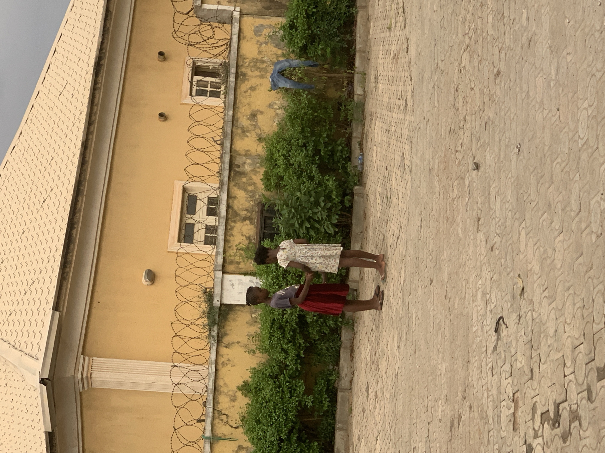 Two young Black girls stand on a patterned-paved ground, likely interlocking concrete pavers, in front of a yellow building with windows and a security barrier of coiled barbed wire. The building also features green shrubbery at its base. One girl wears a red dress, and the other wears a dress with a floral pattern. The image was taken in daylight under an overcast sky. Given the appearance of the barbed wire and the architecture, it is possible the scene is in a residential area of Abuja, Nigeria with heightened security measures. A pair of jeans hangs on the shrubs, drying.