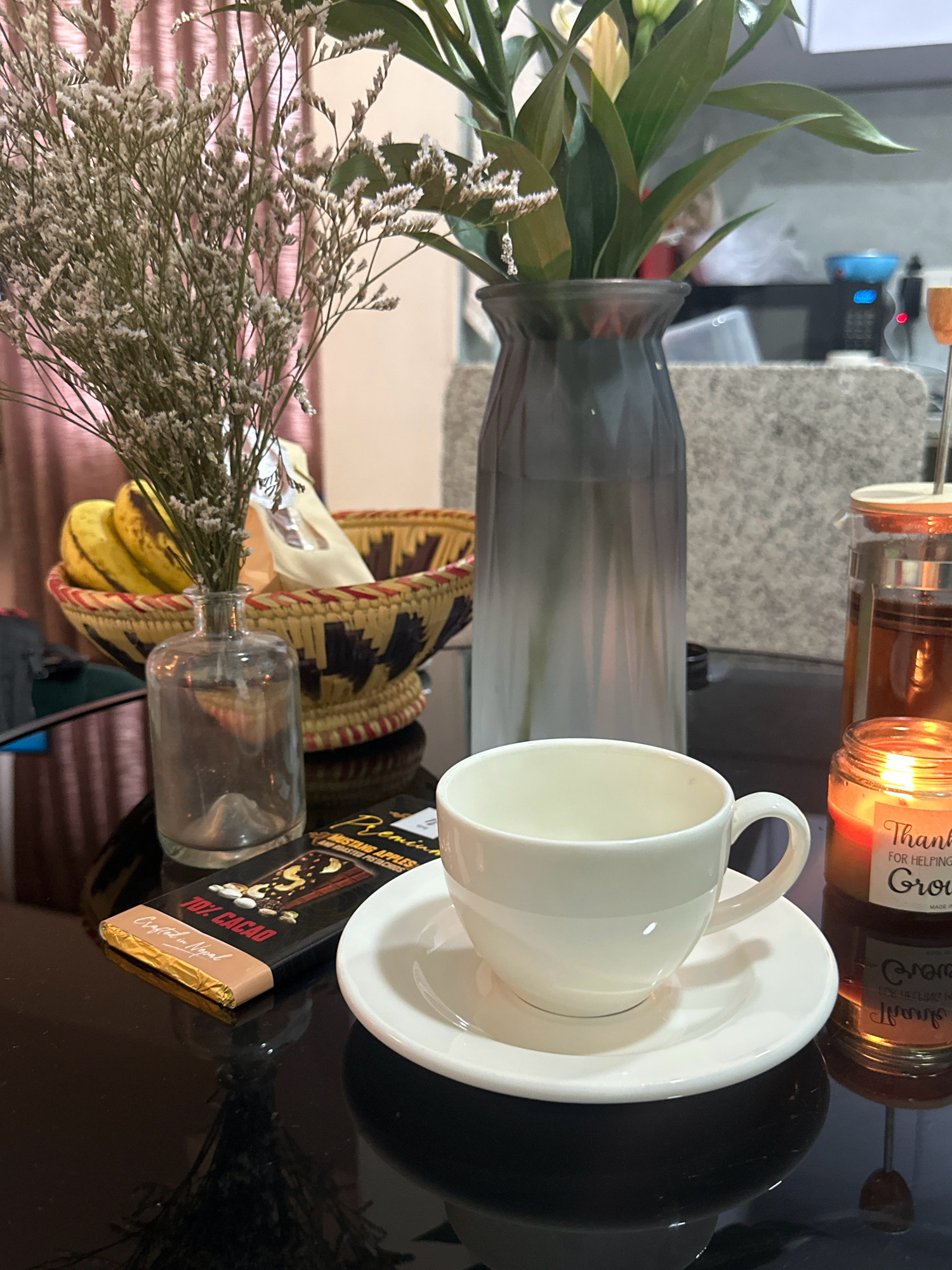 A still life image shows a dark, reflective table with various objects. In the foreground, a white teacup and saucer are positioned to the right. To the left of the teacup is a chocolate bar wrapper that reads "Premium Mustang Apples and Roasted Pistachios 70% Cacao Crafted in Nepal." Behind the chocolate bar is a small clear glass vase containing dried flowers. Further back, a large, grey, gradient vase holds a bouquet of green leaves and some white flowers. To the far right, a lit scented candle in a glass jar reads "Thank You For Helping me Grow." Behind the candle, a glass teapot with amber liquid is visible. In the background, a woven basket filled with bananas and other items sits on the table. The scene appears to be indoors, with soft lighting.