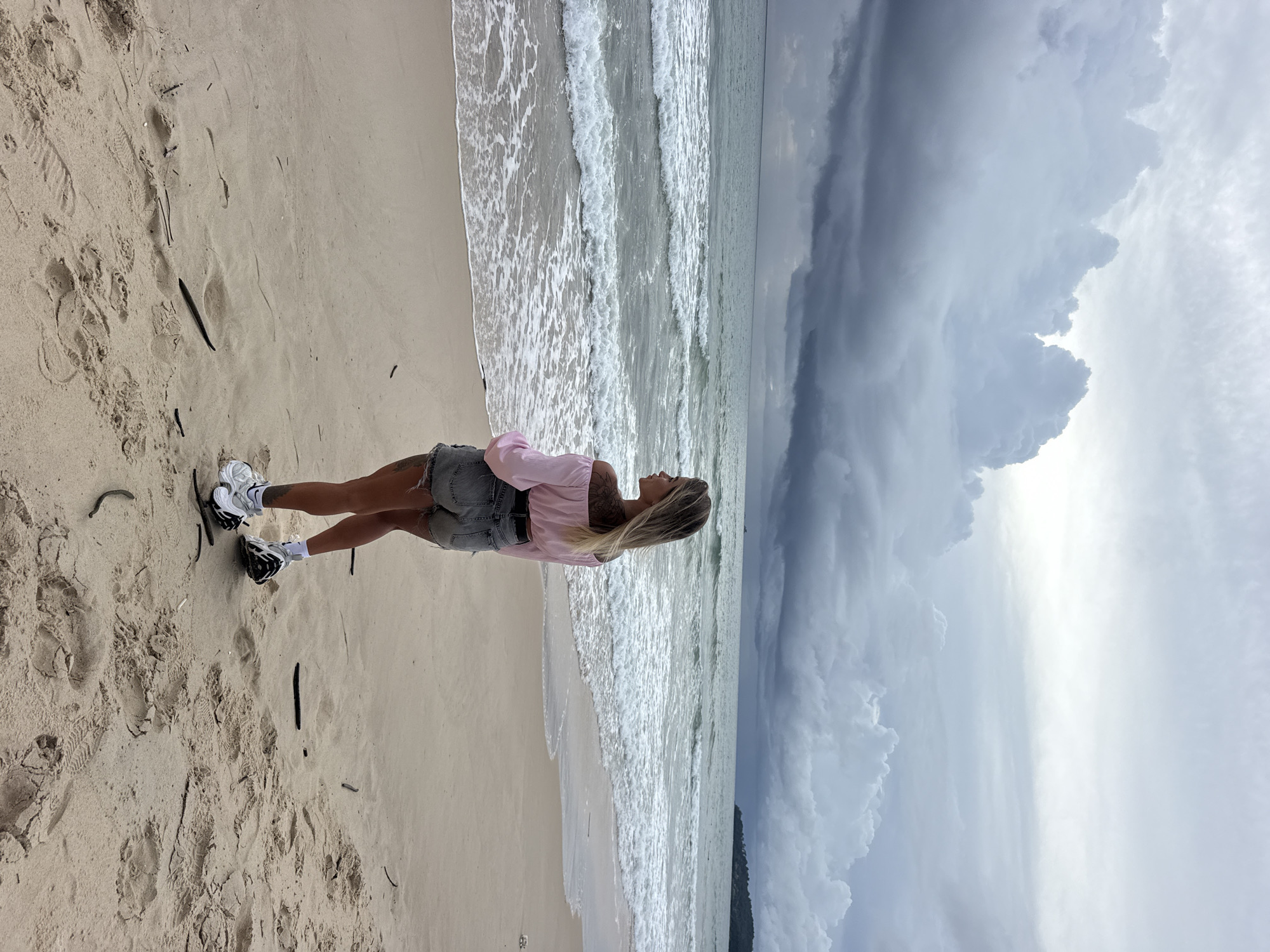A person stands on a sandy beach, facing right, with their arms crossed. They are wearing a pink long-sleeved shirt and denim shorts. The person has blonde hair and tattoos visible on their arm. They are wearing white and black athletic shoes. The scene depicts a beach with waves rolling in. The sky is dramatic, with dark, cloudy formations dominating the upper right portion of the image, suggesting impending rain or a storm. The lighting indicates it could be late afternoon or early evening. The location is Choeng Thale, Thailand, and the overall mood appears contemplative.
