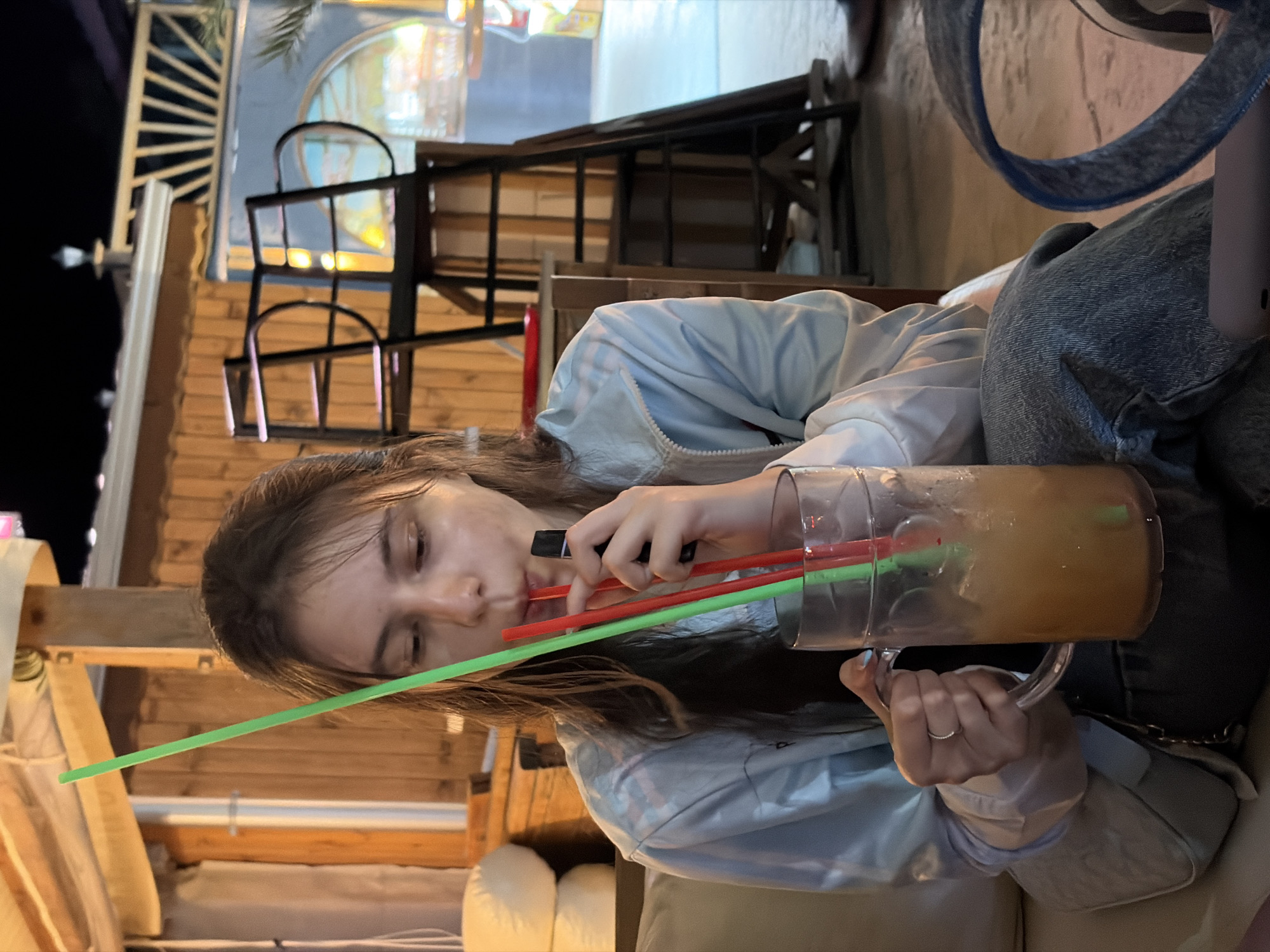 A young woman is sitting at a table, holding a large glass of orange-colored drink with ice and two straws, one green and one red. She is also holding a black vape device. She is wearing a light blue jacket and denim jeans. The background shows a wooden structure with a roof, possibly an outdoor seating area or a shack. Further in the background, there is a metal structure that resembles a ladder or scaffolding, and a circular window with a colorful illuminated scene behind it. The scene appears to be at night, as indicated by the darkness and the artificial lighting. The location is in Varna, Bulgaria.
