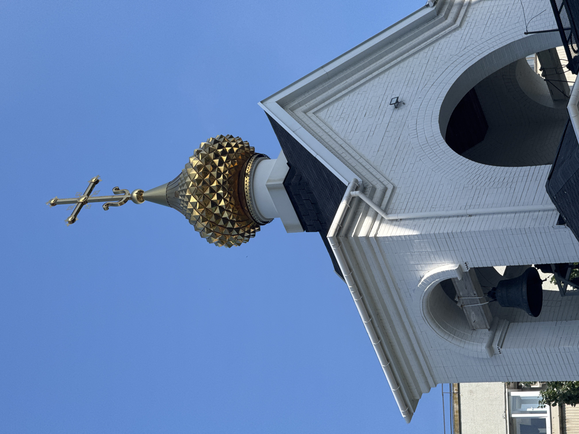 The image shows a portion of a church tower in Unknown, Unknown, viewed from a low angle. The tower features a golden onion dome with a cross on top, a white brick structure with arched openings, and a black roof. A bell hangs within one of the arched openings. The sky is blue and clear.