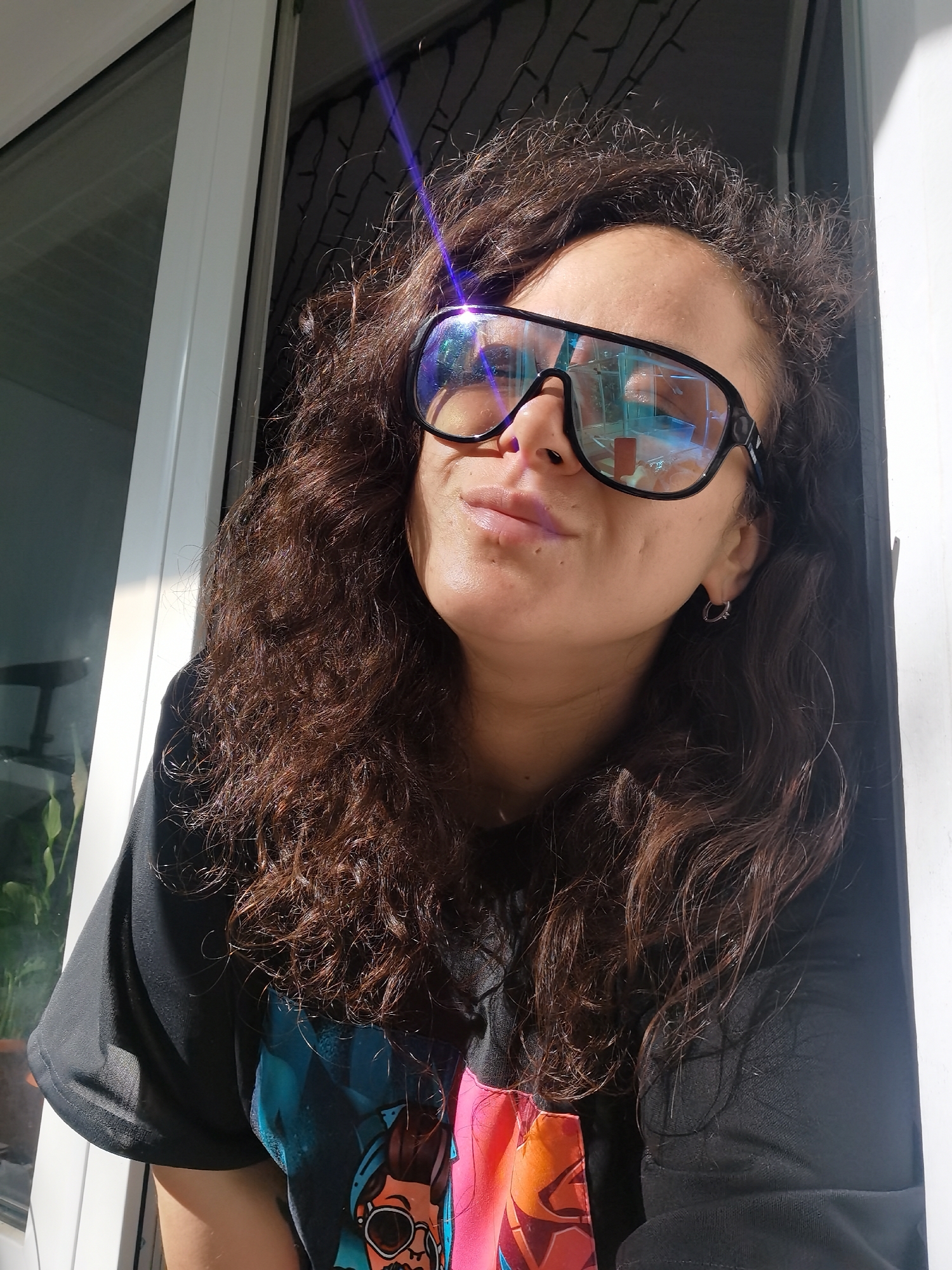 A close-up, eye-level, outdoor medium shot shows a young woman with dark curly hair and brown eyes wearing large, dark-rimmed sunglasses. She is looking slightly down and to her left, with her head tilted. Her lips are slightly parted, and she has a faint smile. She is wearing a black t-shirt with a colorful graphic on it. The background is a white door frame and a window, with sunlight casting shadows on her face and hair. The image is taken in Chaikovsky, Russia.