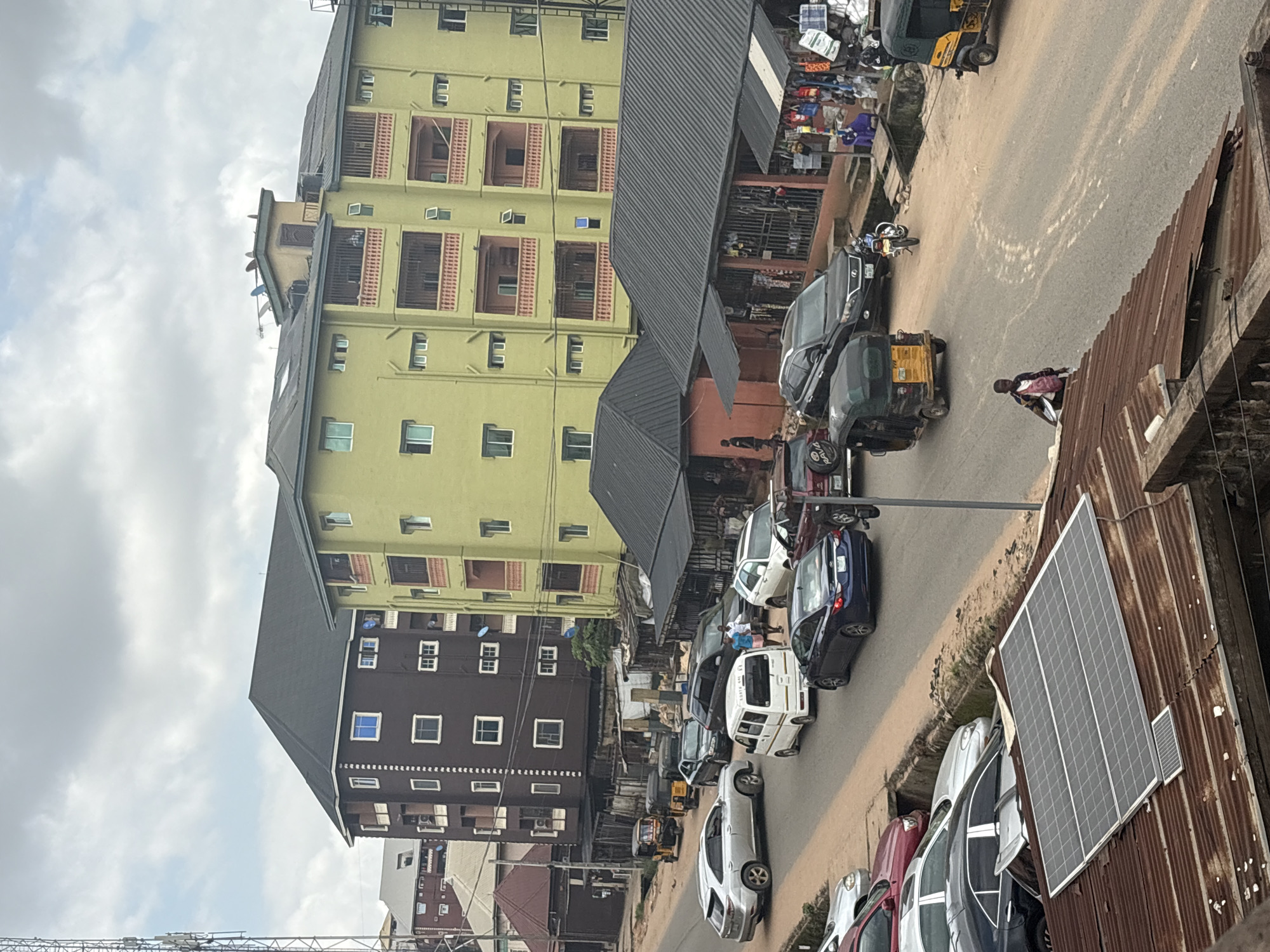 The image captures a street scene in Nkpor, Nigeria, under an overcast sky. Multiple vehicles, including cars, a white van, and several three-wheeled auto-rickshaws, are present on a paved road. Some vehicles are moving, while others appear to be parked or stopped in traffic. Several buildings line the street, with commercial structures featuring awnings and displays of goods along the lower levels. The foreground shows a corrugated metal roof with two solar panels. People are visible on the street and near the buildings, some appearing to be pedestrians and others potentially involved in commerce. The overall impression is that of a busy, urban environment with mixed commercial and residential activity.