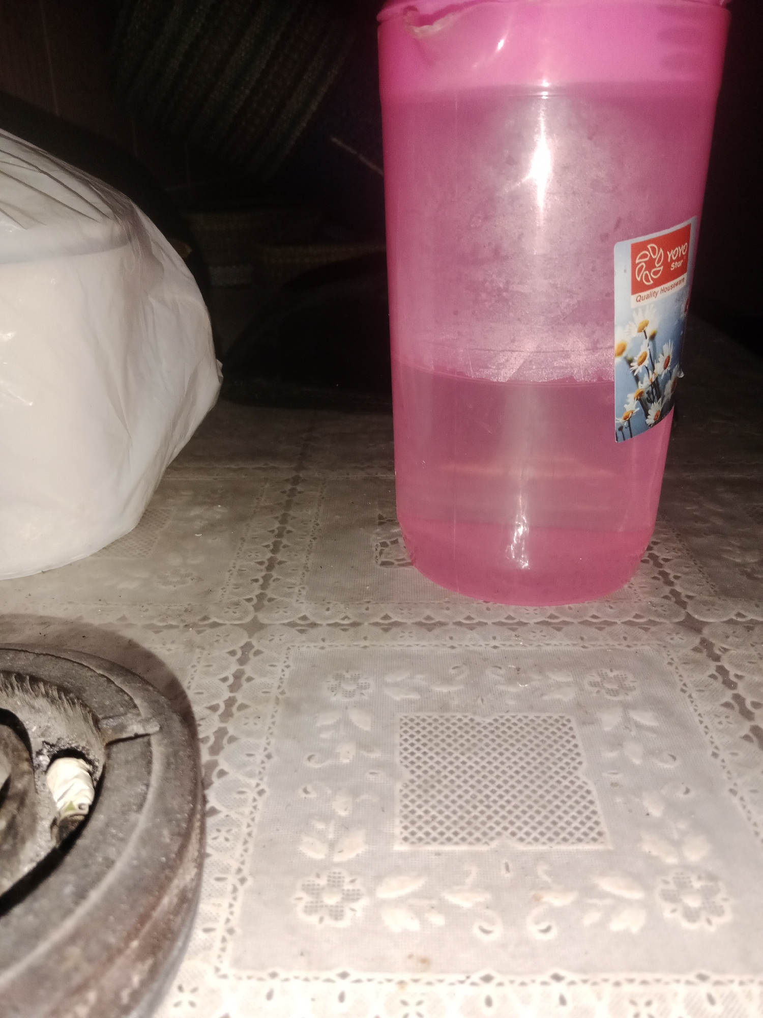 The image depicts a close-up view of a pink plastic cup filled with a pale pink liquid, placed on a white tablecloth with a floral pattern. A white plastic-wrapped object is visible to the left of the cup. In the foreground, a portion of what appears to be a stove element with a spark plug is present. The background is dark and indistinct, suggesting an indoor setting. There are no people or animals visible in the image. The lighting is dim, creating shadows and highlighting the texture of the tablecloth and the condensation on the cup.