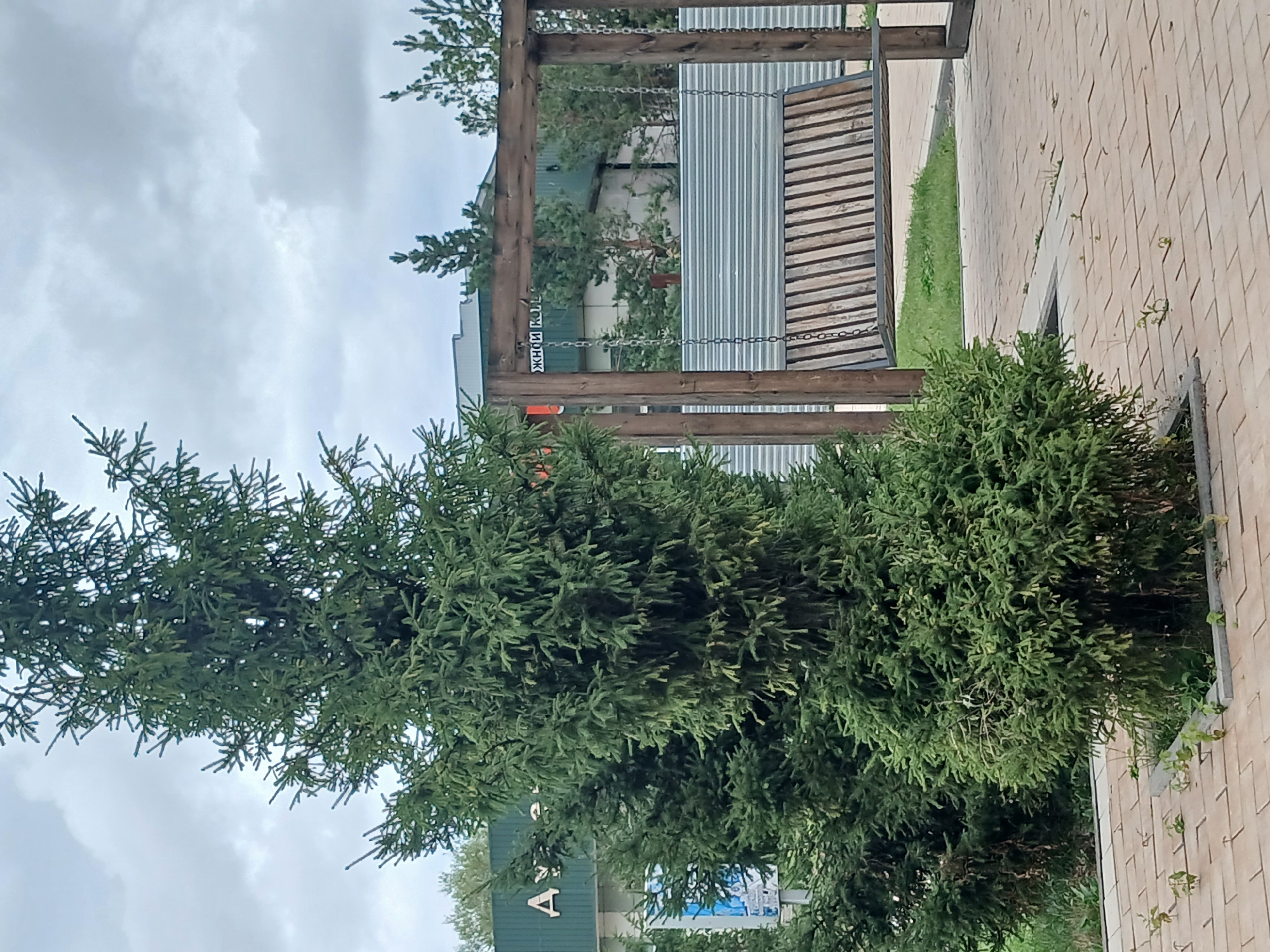 In the city of Shchuchinsk, Kazakhstan, a wooden swing set with a slatted seat hangs from metal chains suspended by a wooden frame. The swing is positioned on a paved area made of light-colored rectangular bricks. To the left of the swing, a tall evergreen tree stands prominently. In the background, a green building with white lettering is visible, partially obscured by trees and a corrugated metal fence. The sky is overcast with grey clouds.