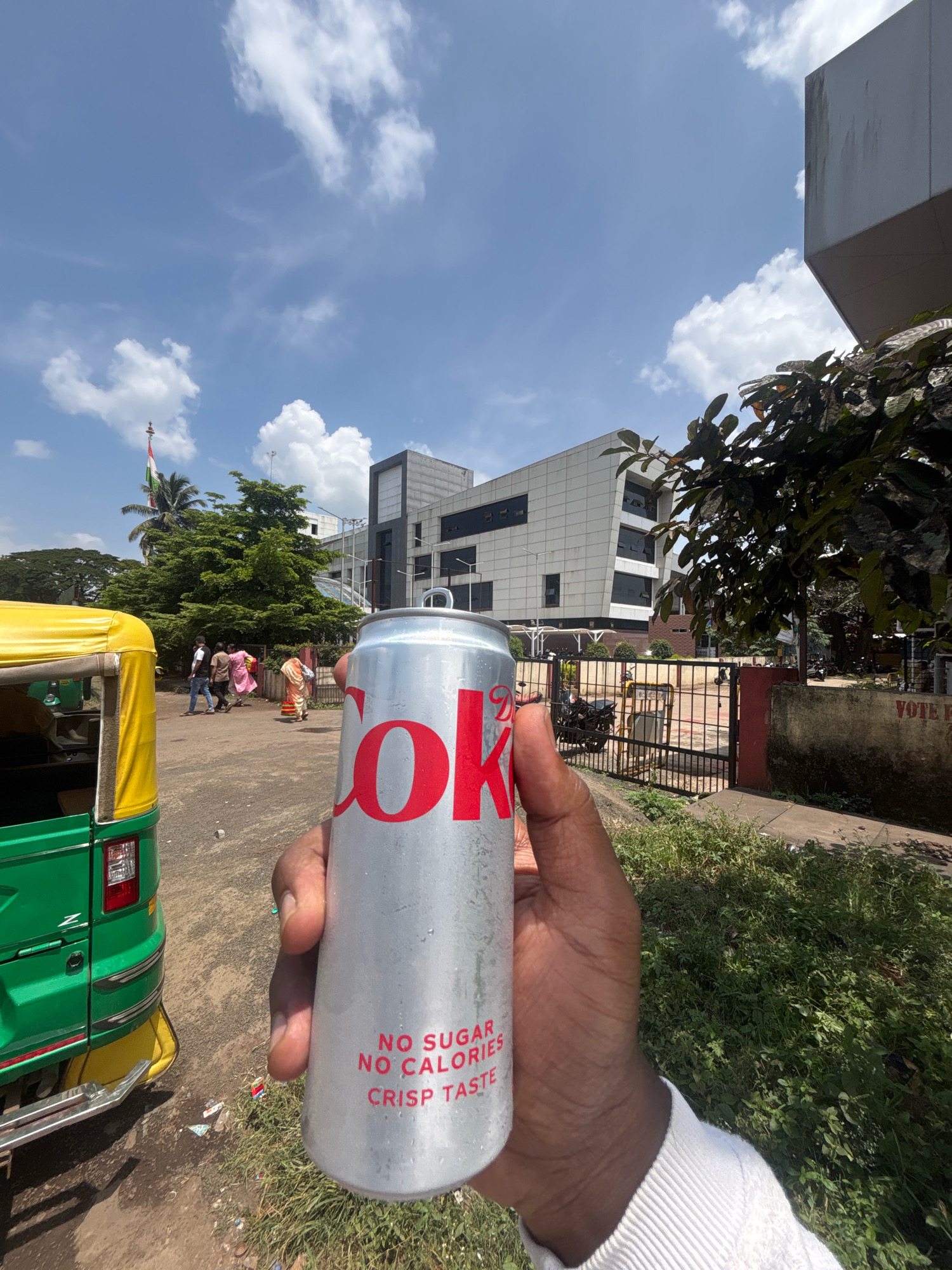 A hand holds a silver can of Coca-Cola Zero Sugar in the foreground, with its distinctive red logo visible. The text "NO SUGAR NO CALORIES CRISP TASTE" is also present on the can. In the background, a green and yellow auto-rickshaw is parked on the left side of the frame. A street scene unfolds behind it, featuring people walking, lush green trees, and a modern white building. An Indian flag flutters on a flagpole to the left of the building, suggesting the location is Belagavi, India, potentially near a government or public institution. The sky is blue with scattered white clouds, indicating a sunny day. The overall scene depicts an urban environment with a touch of everyday life.