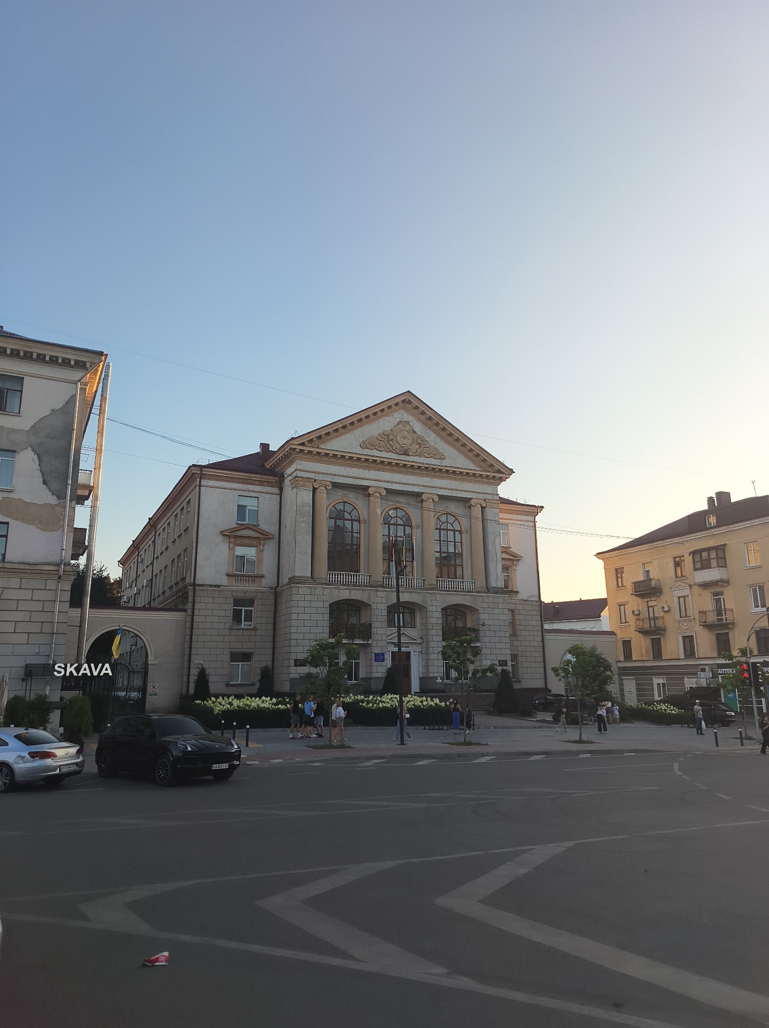 In Ternopil, Ukraine, a wide-angle, eye-level shot captures the facade of a grand building with classical architectural elements. The building features a prominent triangular pediment adorned with intricate carvings, supported by large columns. Large arched windows line the upper floors. In front of the building, a well-maintained area with manicured hedges and trees is visible. A street with cars and pedestrian traffic runs in the foreground. The sky is a clear, pale blue, indicating daytime, likely late afternoon or early evening due to the soft lighting. The overall impression is one of civic architecture and urban life.