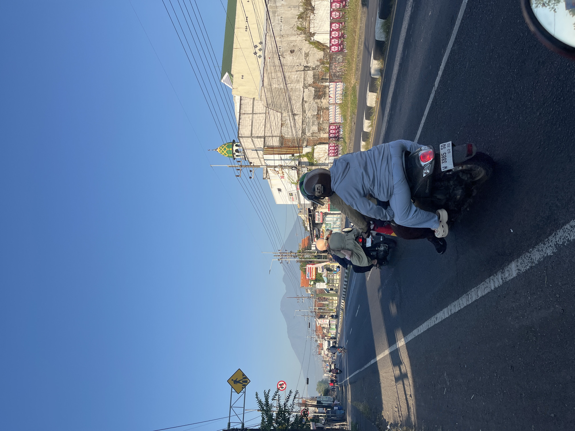 This image captures a street scene in Siring, Indonesia, likely during the day. In the foreground, two individuals are riding on a motorcycle, with the rider wearing a helmet and the passenger behind them also wearing a helmet. The passenger is dressed in a light blue hooded sweatshirt and light blue pants. The motorcycle has a license plate reading "W 3885 SQ". The road is paved and marked with white lines.
In the background, other motorcycles and a car are visible on the road. Buildings line the street, some appearing commercial with signage. One building features a green and yellow domed structure, possibly part of a mosque. A prominent mountain range is visible in the distance under a clear blue sky. Traffic signs, including a pedestrian crossing sign and a prohibitory sign, are also present. The environment appears to be an urban or semi-urban area with clear weather.