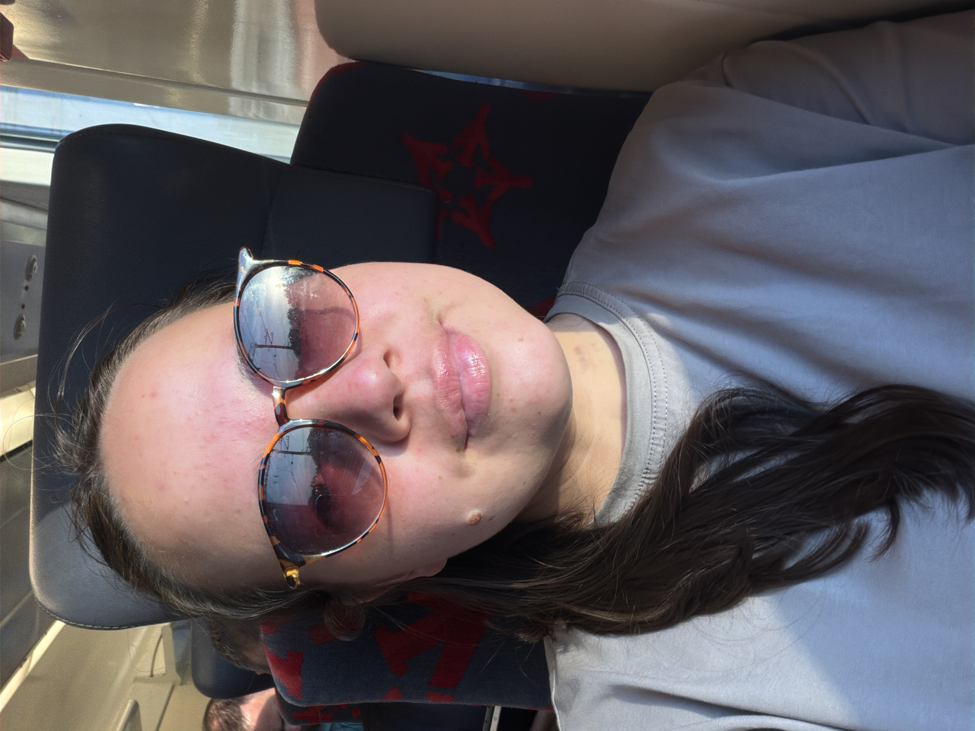 A woman with fair skin and dark hair lies on a train seat wearing round sunglasses. The seat is gray with red star-shaped patterns. Outside the train window, reflections of an unidentified landscape are visible in the sunglasses lenses. There is no evidence in the image to place the location in Moscow, Russia.