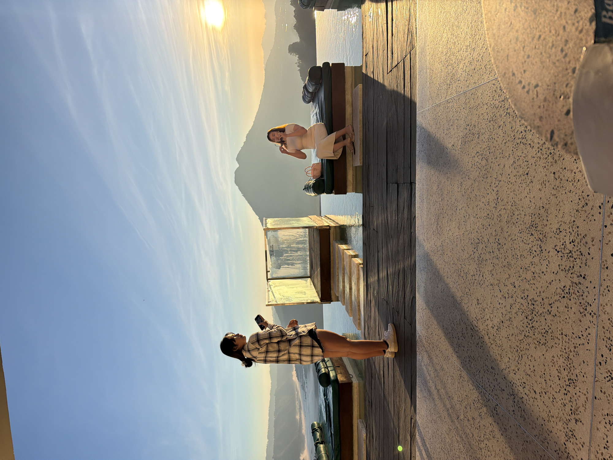 The image depicts an outdoor scenic overlook in Kintamani, Indonesia, at sunrise or sunset, characterized by warm, golden light and long shadows. Two women are present on a multi-level deck overlooking an infinity pool and a mountainous landscape.
In the foreground, a textured, speckled light-colored concrete floor transitions to a darker wooden plank deck. A small, bright green spherical object rests on the wooden deck near the left woman's feet.
On the left, a woman stands in profile, facing right. She has dark hair tied back and wears a plaid long-sleeved shirt (white, black, and beige checks) over dark shorts and white sneakers. She holds a dark smartphone in both hands at chest height, looking at its screen. A translucent glass barrier with metal framing stands behind her, featuring water on its surface.
On the right, a second woman is seated on a dark green, cushioned lounge bed that is partially submerged in the infinity pool. She faces left, angled slightly towards the viewer. She has light brown hair and wears a light-colored (possibly beige or light pink) short-sleeved top and a matching long skirt. Her right hand is raised, holding a pair of sunglasses near her mouth/face. A small, light pink handbag rests beside her on the lounge bed.
The infinity pool stretches across the midground, containing several such lounge beds and light-colored stepping stones leading into the water on the left.
In the background, a large, prominent mountain (likely Mount Batur) rises above a layer of mist or low-lying clouds that fills the valley below. The mountain is silhouetted against the bright, transitioning sky. The sun, a bright golden disk, is visible in the upper right quadrant, casting strong illumination and long shadows across the scene. The sky above is clear blue with faint, wispy clouds.