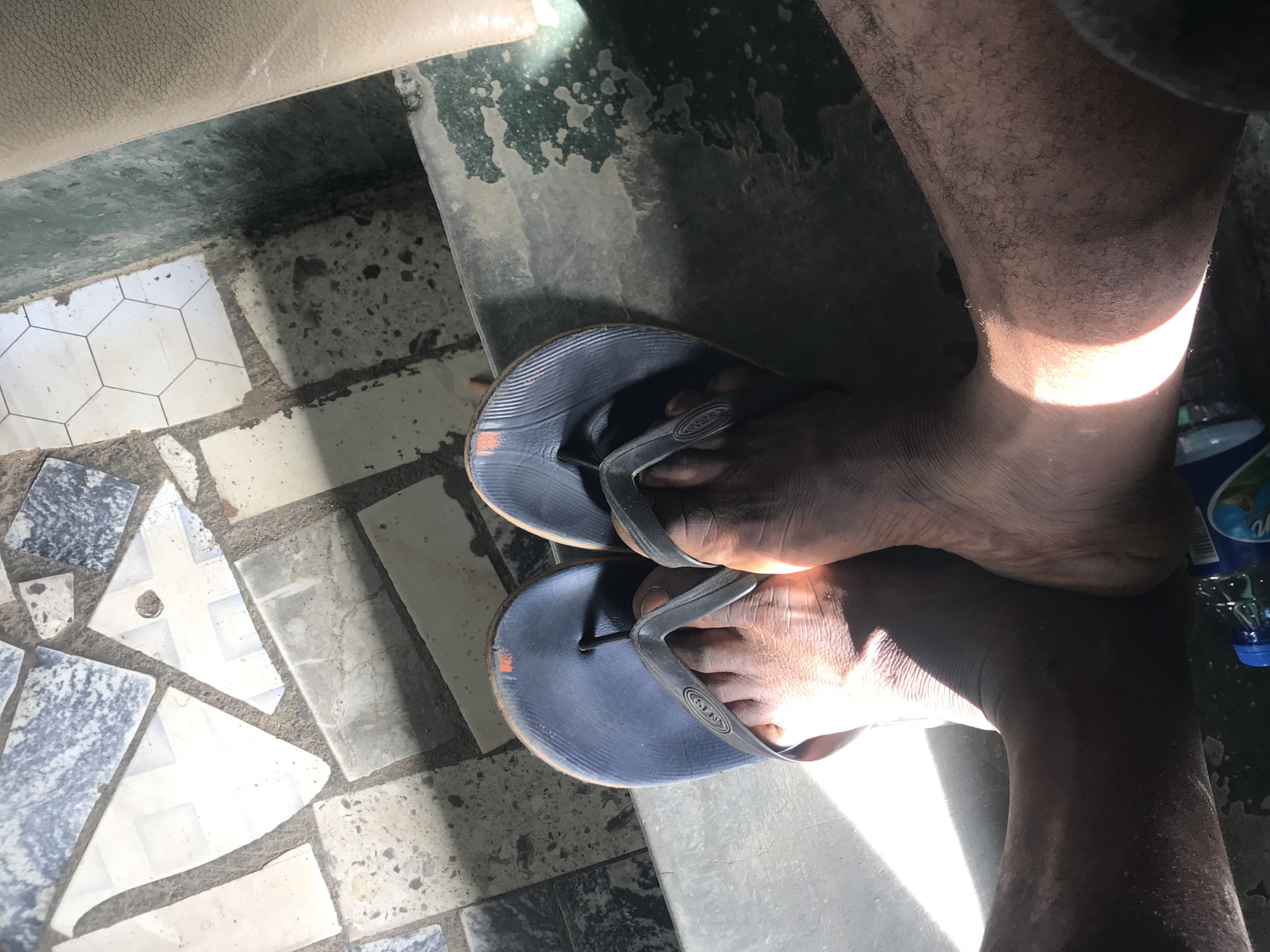 In New Stadium, Nigeria, a pair of dark blue flip-flops rests on a concrete ledge. One flip-flop, worn on the left foot, displays a small orange mark on its sole. Sunlight casts sharp shadows across the scene, illuminating the textured soles of the flip-flops and the skin of the feet. The background reveals a tiled floor composed of various geometric shapes and colors, interspersed with grout lines and some debris. To the right, a portion of a beige upholstered surface is visible, possibly a car seat or furniture. In the lower foreground, a clear plastic bottle with a blue cap, partially filled with liquid and a label, lies on the ground. The overall environment appears to be an outdoor or semi-outdoor space with varied textures and lighting conditions.