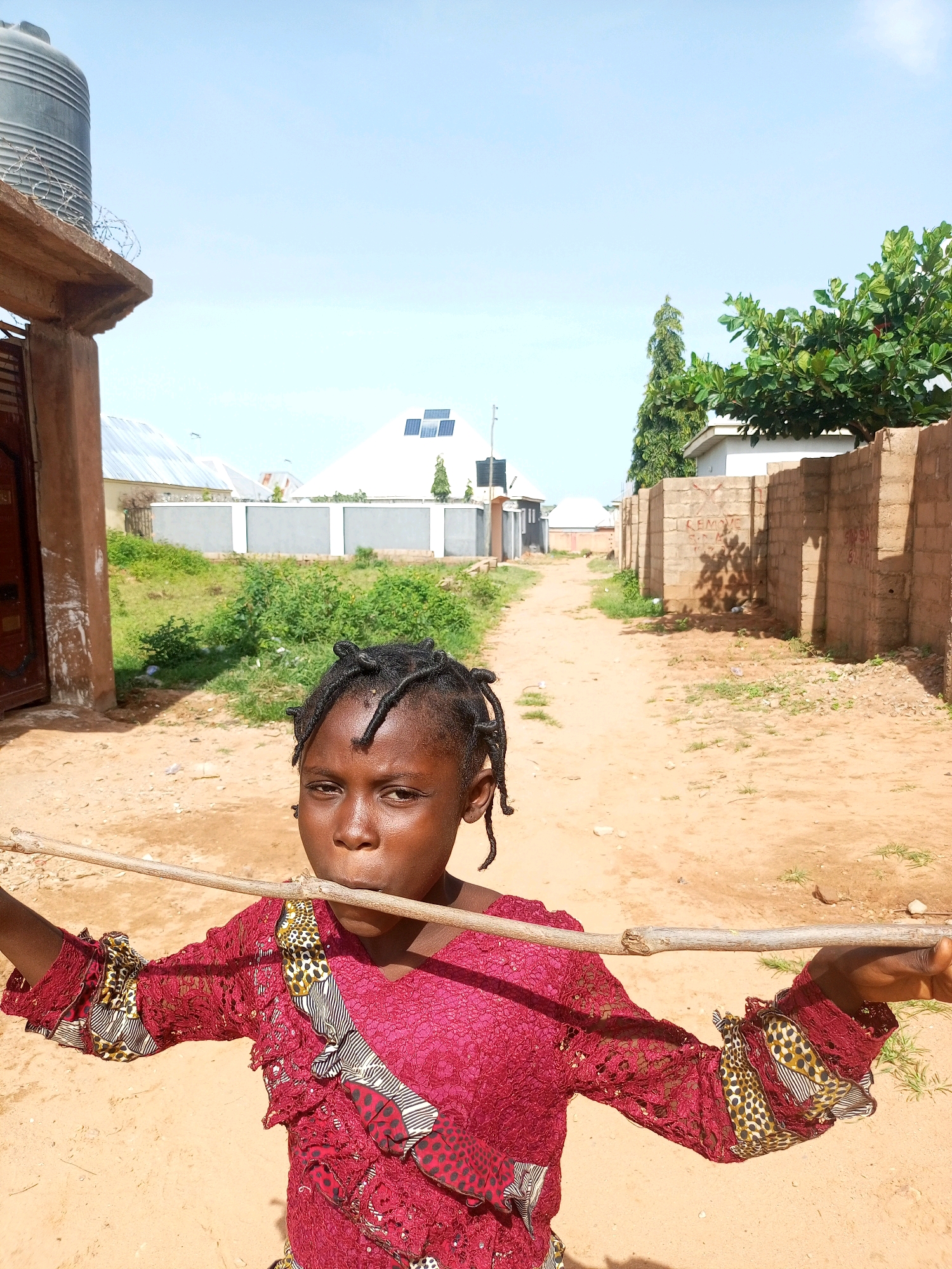 A young Black girl with braided hair stands in a dirt alleyway in Gombe, Nigeria. She is wearing a bright red, lacy dress with patterned accents on the sleeves and bodice. She holds a thin wooden stick horizontally in front of her mouth, appearing to be playing with it. Behind her, the alleyway is lined with brick walls and some vegetation. To her left, a brown door is visible on a building, and a large grey water tank sits atop a concrete structure. Further down the alley, buildings with metallic roofs and solar panels can be seen under a clear blue sky. The overall scene depicts a street in a residential area of Gombe.