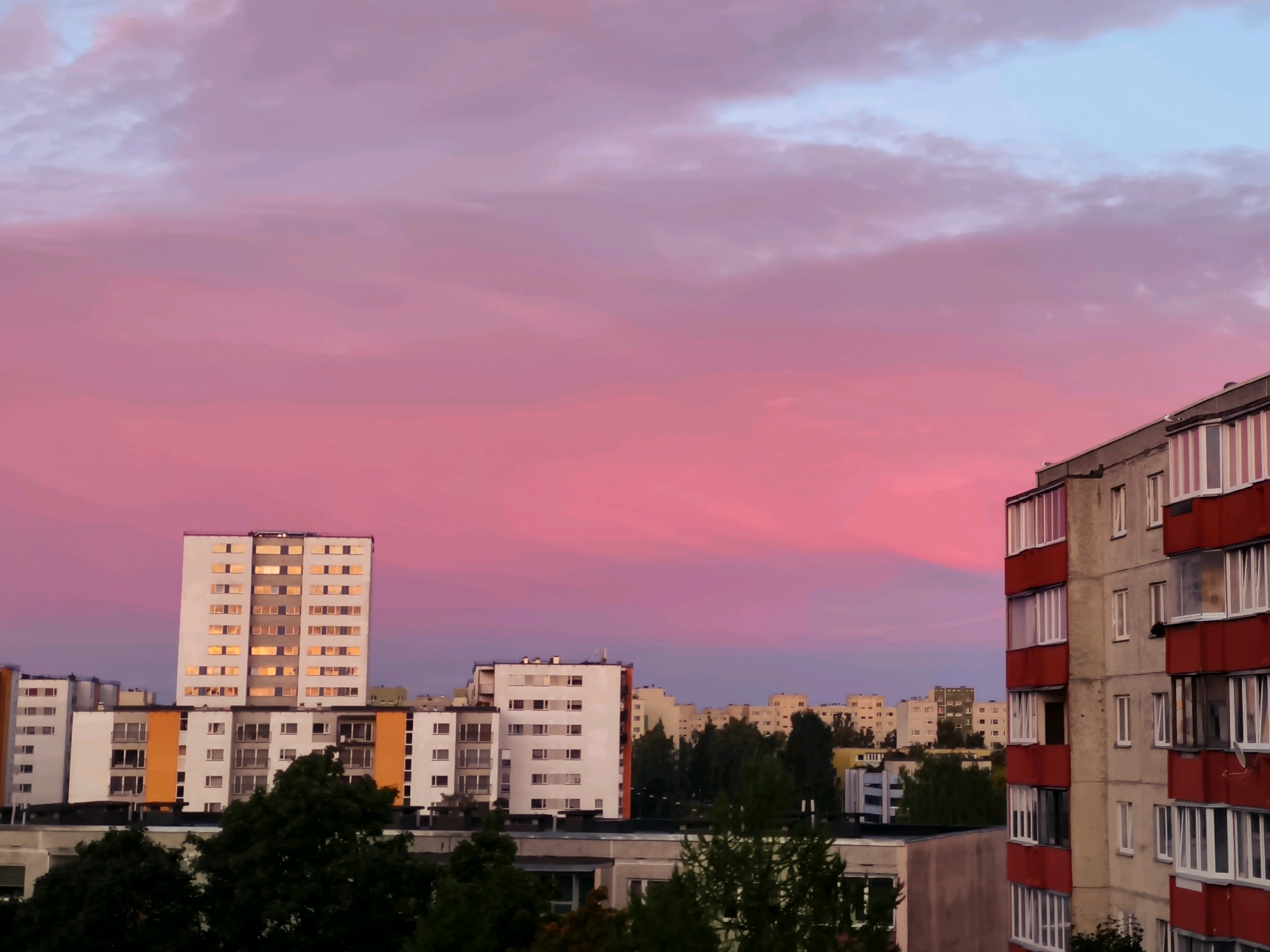 The image captures a twilight scene in the Lasnamäe district of Tallinn, Estonia. A vibrant pink and purple sky dominates the upper portion of the frame, with soft clouds suggesting the fading light. In the foreground, several multi-story apartment buildings are visible.
The most prominent building on the left is a tall, white residential block with numerous windows, many of which reflect the warm hues of the sunset. To its right, a shorter building features orange and white panels and a similar pattern of windows. Further to the right, a closer apartment building displays a concrete facade with distinctive red trim around its balconies and window frames.
In the lower part of the image, trees and the roofs of other buildings suggest a dense urban environment. The overall impression is one of a residential area at dusk, illuminated by the dramatic colors of the sky. No people are visible in the frame.