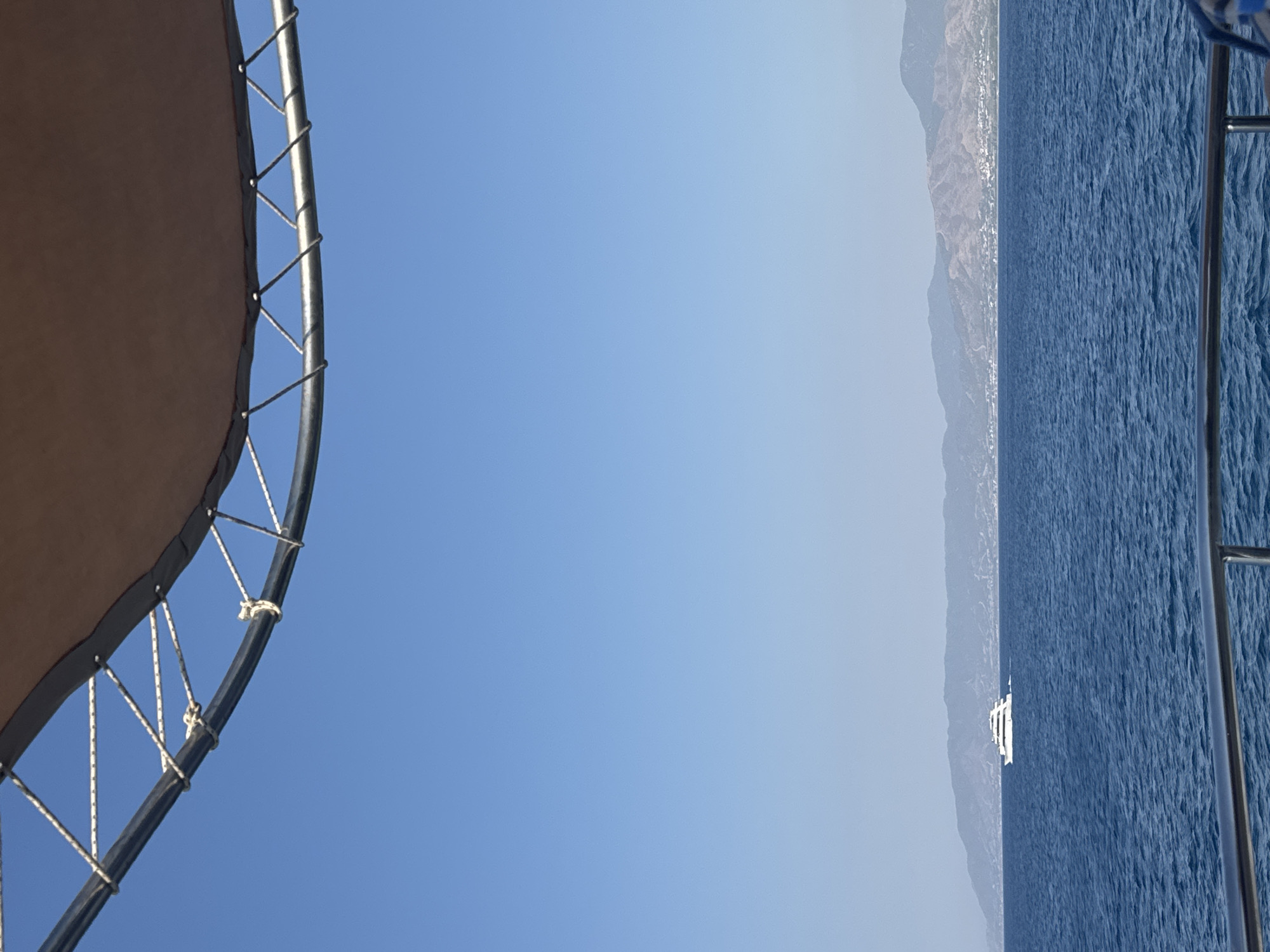 A white yacht is visible on a deep blue sea, with a distant mountainous coastline in the background. The coastline appears to be populated with buildings. The foreground shows part of a boat's railing and a canopy structure made of metal tubing and brown fabric. The sky is a clear, bright blue. The scene is outdoors on a sunny day.