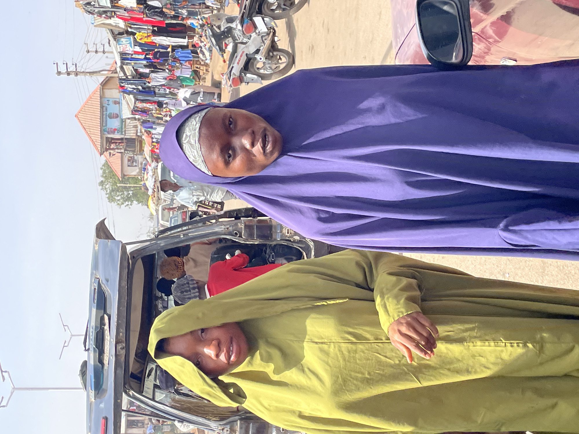 The image presents a vibrant daytime street scene in Bukuru, Nigeria, with two women prominent in the foreground. The woman on the right wears a purple hijab over a patterned white headscarf, looking towards the camera with her mouth slightly ajar. Next to her, on the left, a second woman wears a lime green hooded garment, also looking towards the camera with a subtle smile. Her left hand rests on her clothing.
They appear to be situated within or beside a vehicle, with a partial view of a blue vehicle's rear visible in the mid-ground. The background is a bustling market, featuring numerous stalls overflowing with clothes and various goods. A white motorcycle is parked amidst the market activity. Behind the market stalls, a building with an orange-tiled roof and a light-colored facade is visible. On this building, a blue banner displays some partially legible text and graphics, including what appears to be "4+4" and possibly political campaign imagery. Utility poles and overhead wires crisscross the bright, clear sky. Several other individuals are seen in the background, either walking or inside vehicles, contributing to the lively atmosphere. The scene depicts a sunny day, with strong light casting shadows.