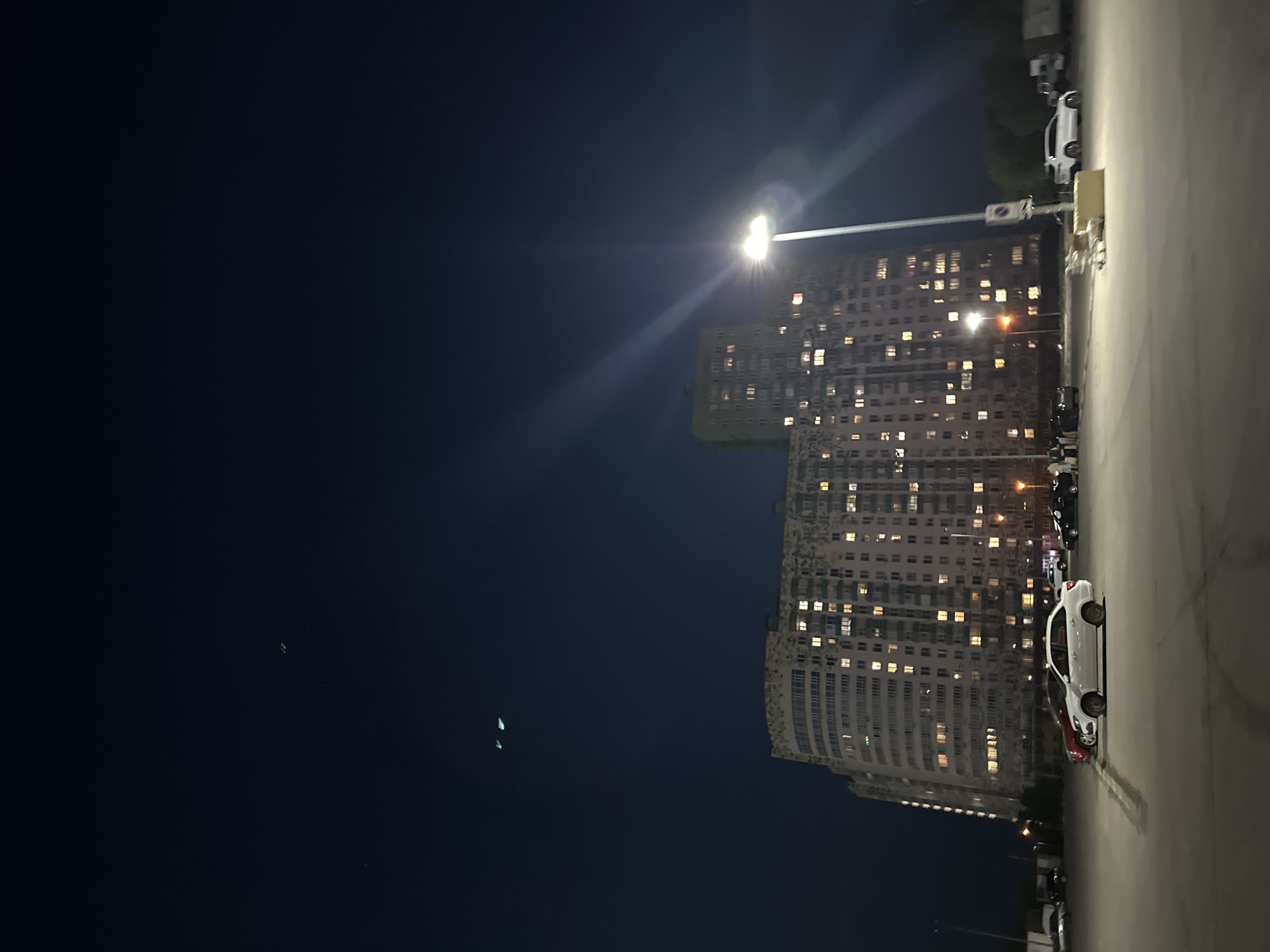 The image shows a nighttime view of a building in Nizhny Novgorod, Russia. The building is tall and rectangular, with many illuminated windows. A street light shines brightly in front of the building. In the foreground is a parking area with several cars. The sky is dark.