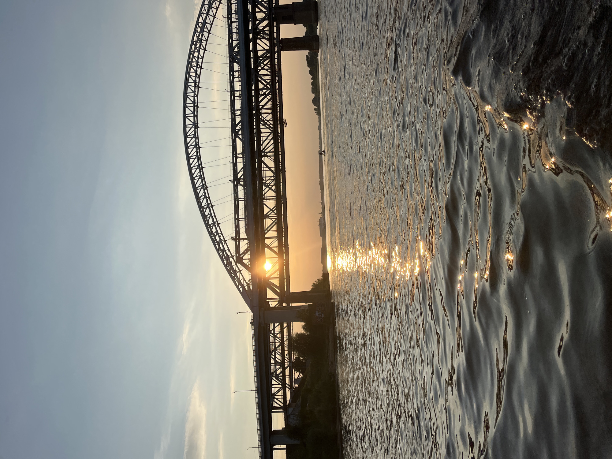 This image captures a sunset over a wide river, likely the Volga River, in Nizhny Novgorod, Russia. A large, multi-tiered bridge dominates the scene, with the setting sun peeking through its structure, casting a golden glow on the water. The bridge appears to be a combination of road and rail, with a distinctive arched metal framework on the upper level and a more utilitarian concrete and steel structure below. The water's surface is rippled, reflecting the sunlight in shimmering patterns. The riverbanks are visible, featuring lush greenery and distant buildings, suggesting an urban or semi-urban setting. The sky is a pale blue with wispy clouds, characteristic of a late summer or early autumn evening. No people are clearly discernible in the image, though there might be small vessels on the water in the distance. The overall impression is one of serene natural beauty juxtaposed with impressive human engineering.
