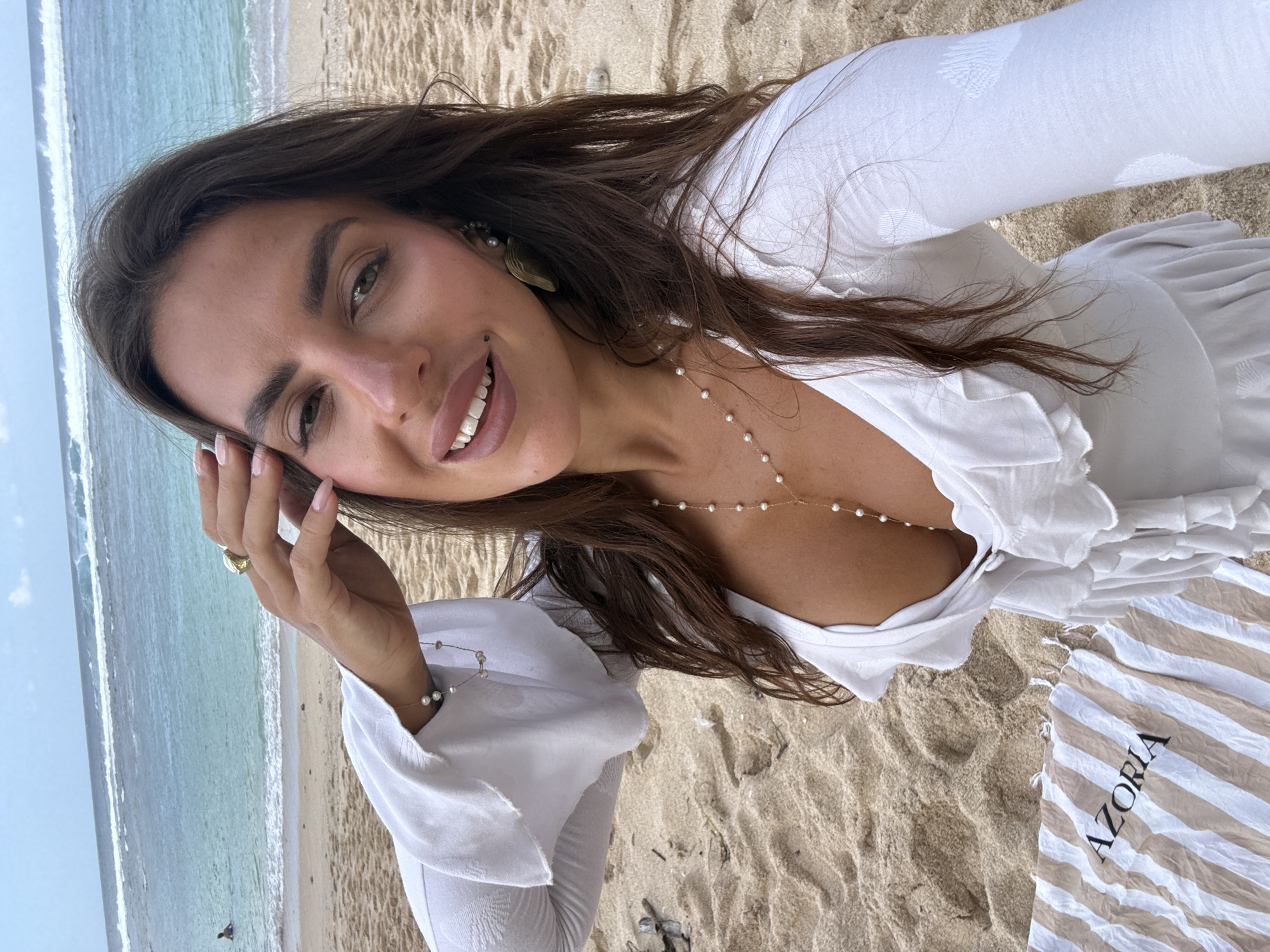 A woman is taking a selfie on a beach in Kuta Selatan, Indonesia. She is wearing a white dress with ruffled details and a pearl necklace. Her hand is touching her hair, and she is smiling. The ocean is in the background, with waves breaking on the shore. There is a towel with the word "AZORIA" on it next to her.