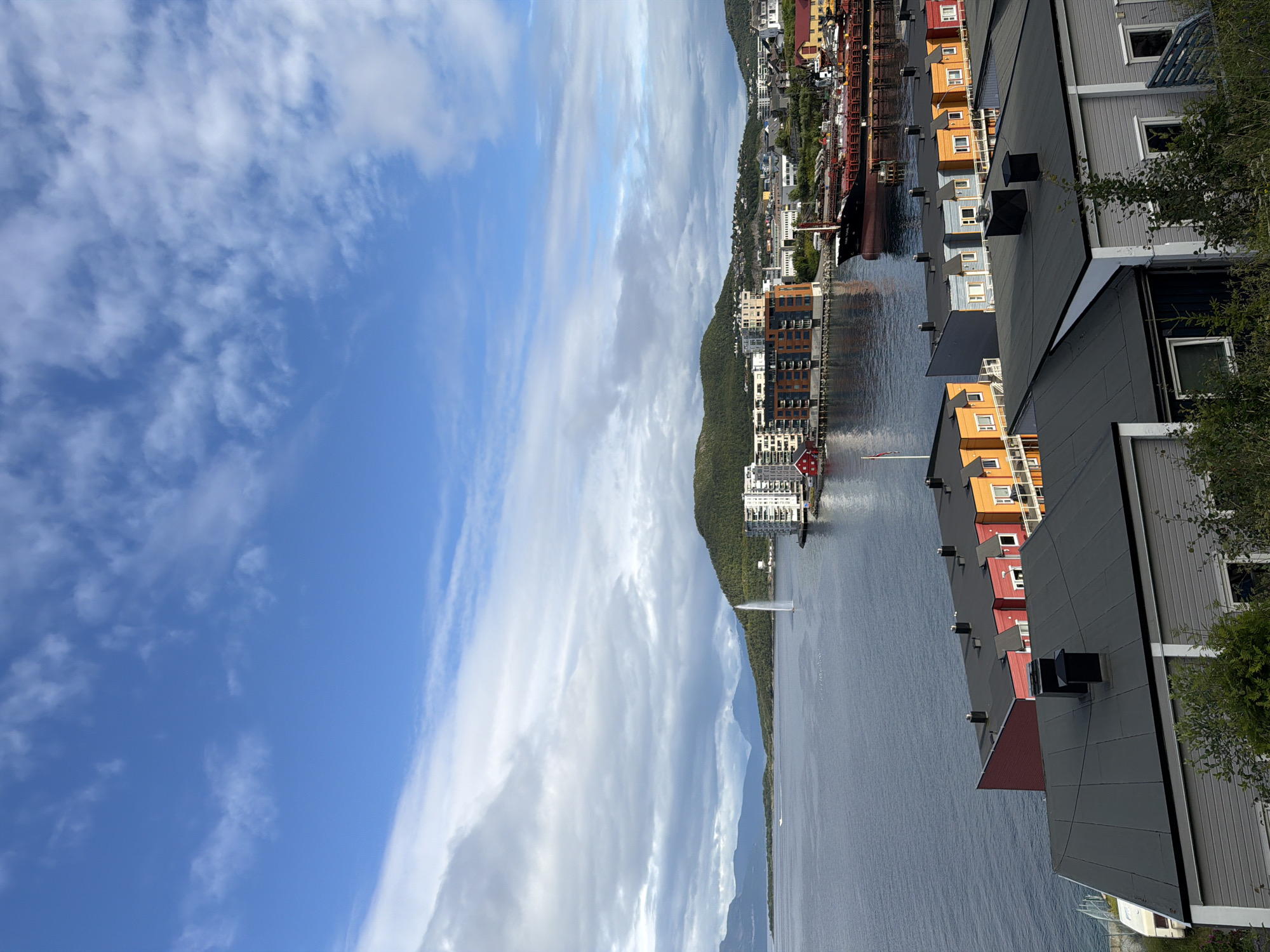 The image shows a view of the waterfront in Harstad, Norway. In the foreground, there are several colorful houses with dark roofs. Further back, along the water, are modern apartment buildings and a large ship docked in the harbor. A water fountain is visible in the bay, and hills covered in trees rise in the background. The sky is blue with scattered clouds.