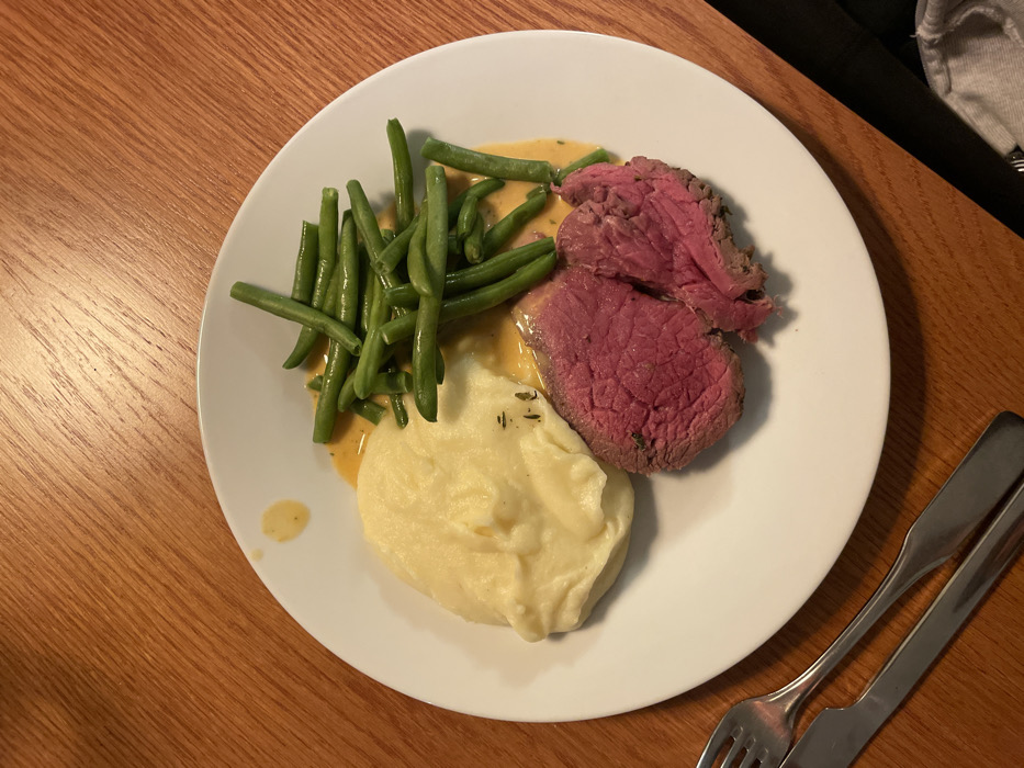 On a white plate sits a meal of steak, mashed potatoes, and green beans. The steak, appearing medium-rare, is sliced and positioned on the right. A scoop of creamy mashed potatoes rests to the left. Fresh green beans are piled high on the upper left, drizzled with a light sauce. A similar sauce is underneath the steak and potatoes. A fork and knife are visible on the lower right, situated on a wooden table.