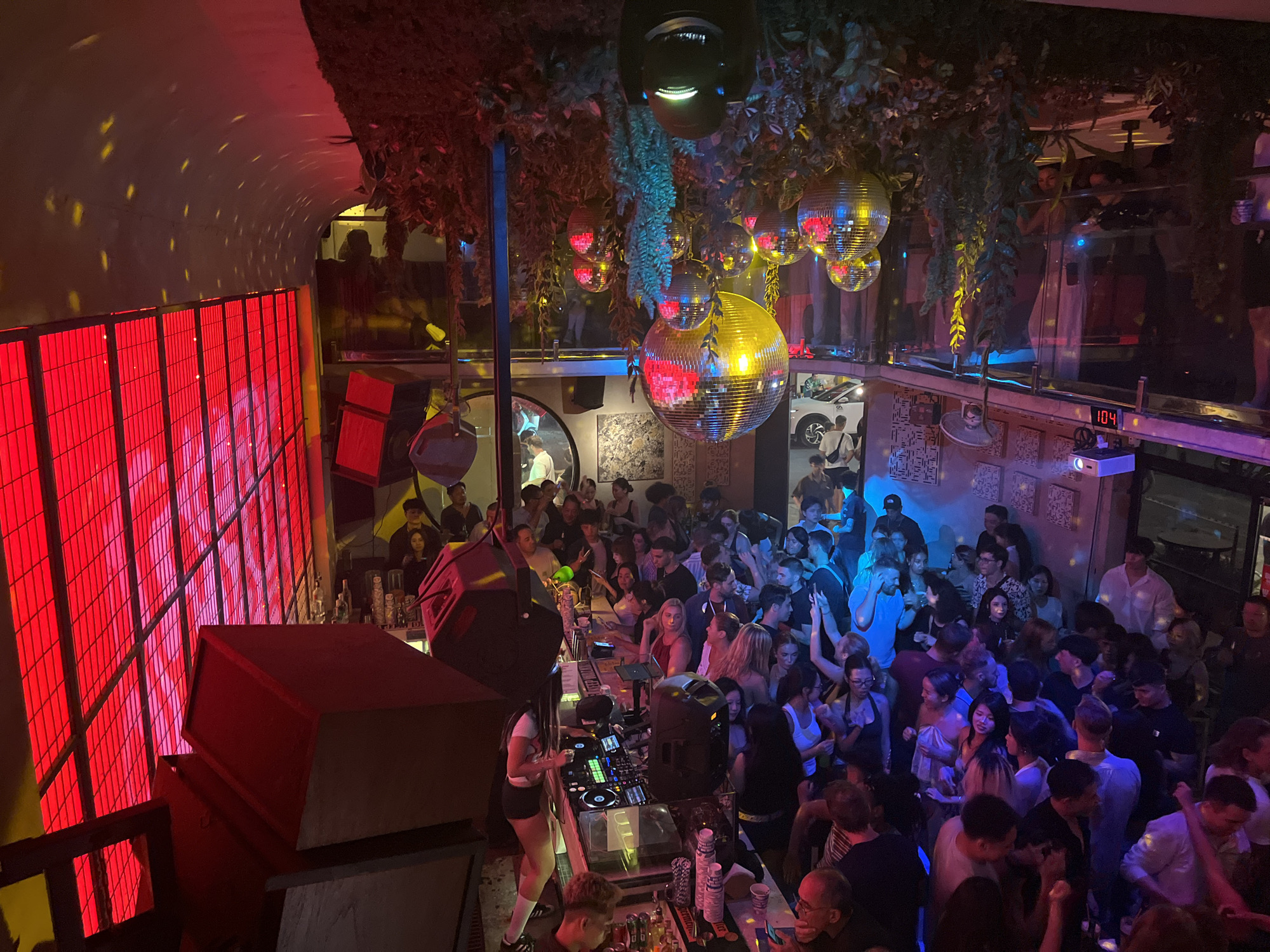 The image shows a crowded nightclub, likely in Đà Nẵng, Vietnam. A DJ is playing music from a booth, with a large crowd of people dancing and socializing in front of the booth. The club has a multi-level layout, with a second level visible above the main floor. The club's decor includes disco balls, greenery hanging from the ceiling, and a red light wall feature. People are drinking and interacting, and the overall atmosphere is lively.