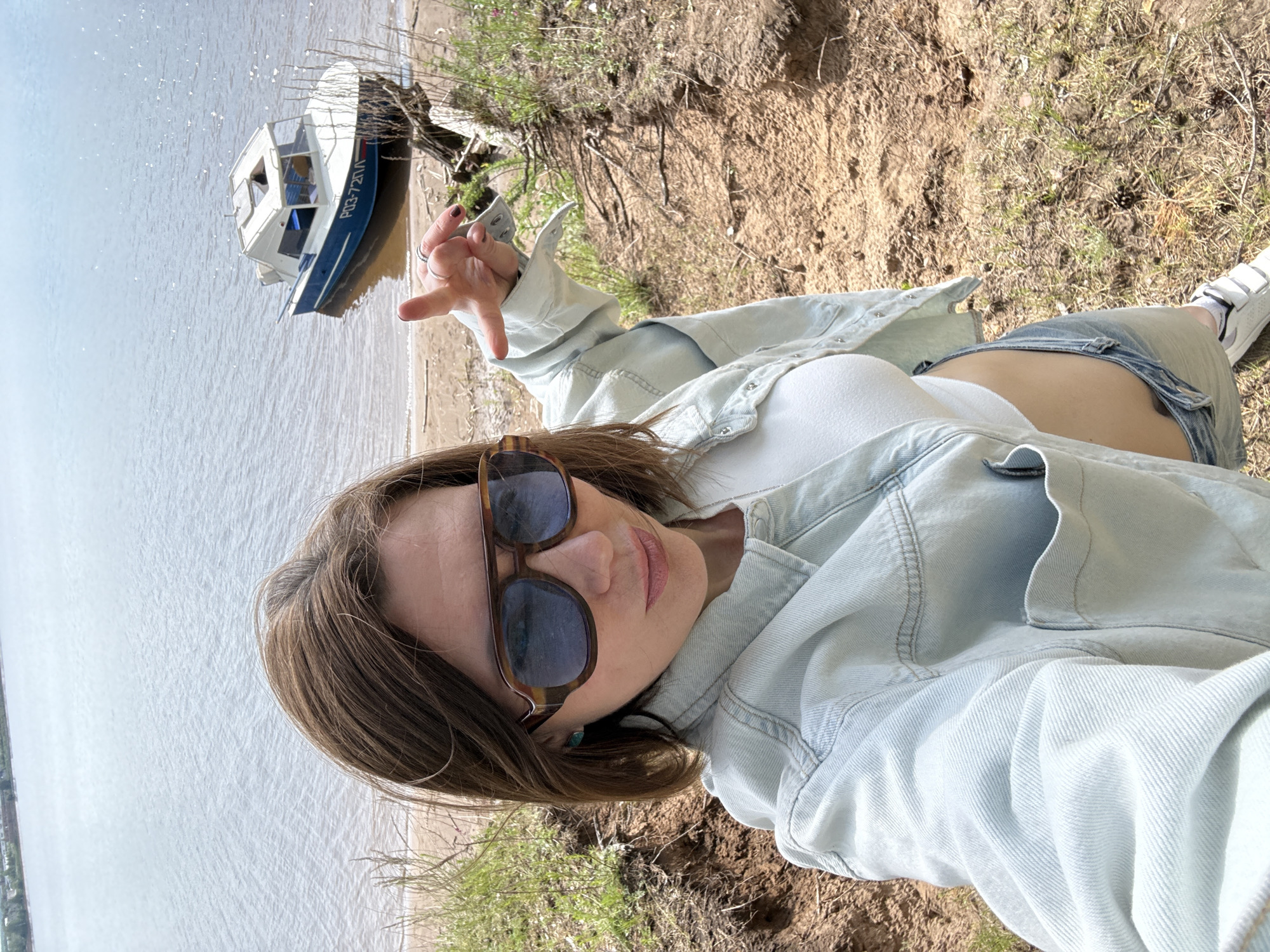 This is an outdoor selfie featuring a woman posing on an eroded dirt bank overlooking a body of water in Chaikovsky, Russia. The woman, with brown hair, is wearing sunglasses, a white crop top that exposes her midriff, a light blue denim jacket, and blue denim shorts. White sneakers are partially visible on her feet. She has rings on her fingers and a small blue or green earring in her right ear. Her expression is neutral and relaxed as she takes the photograph.
Behind her, down the bank and partially submerged in shallow water near a sandy shore, is a small boat. The boat is white with a blue hull and has the text "РОЗ-72ЛК" clearly visible on its side. The water appears calm, and the distant horizon is faintly visible. The bank consists of loose soil with patches of green grass and other sparse vegetation. The scene is brightly lit, indicating a sunny daytime.