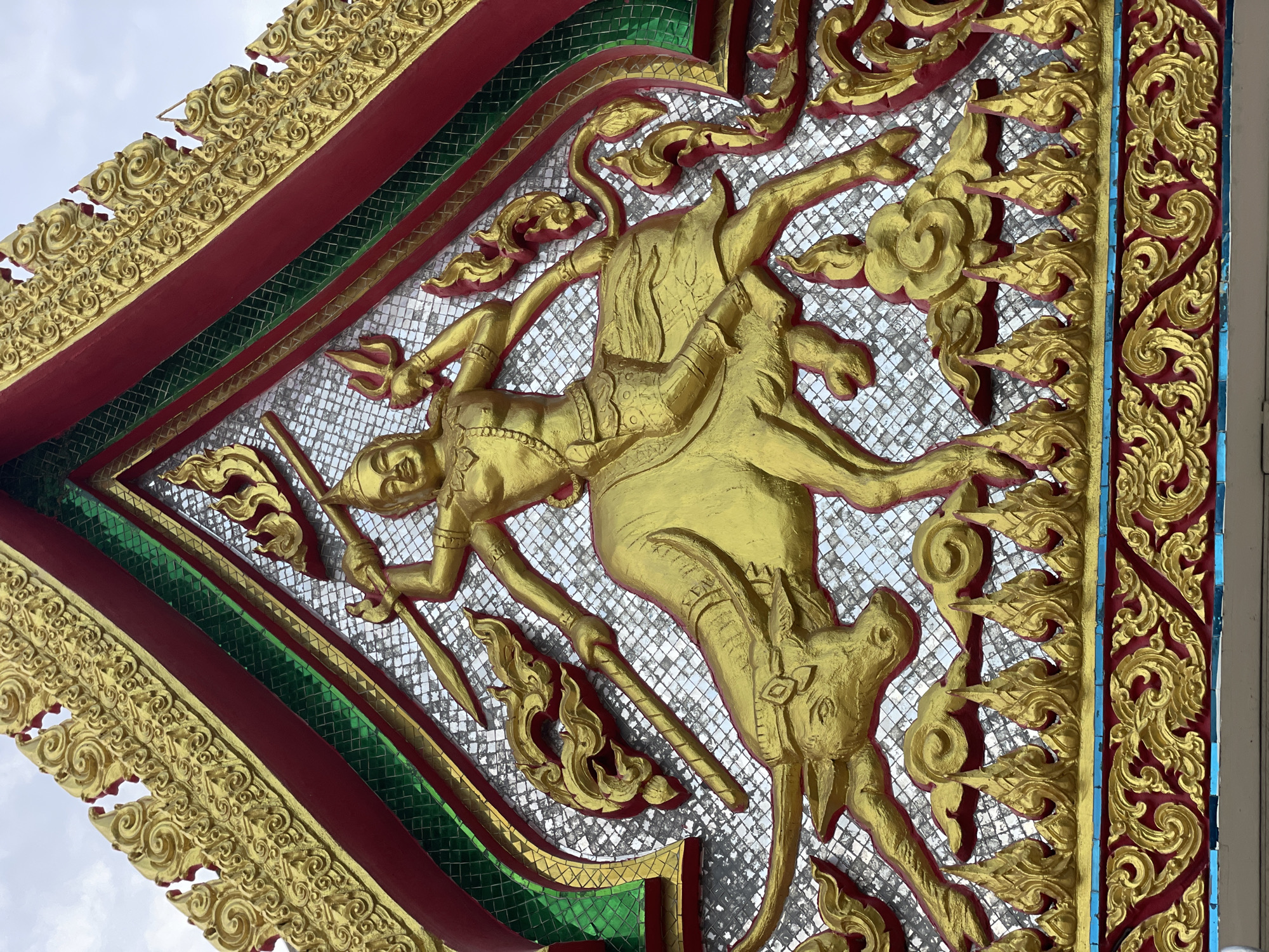 A close-up, low-angle, eye-level outdoor shot showcases an ornate, golden bas-relief sculpture on the facade of a building, likely a temple or a decorative structure, in Pattaya City, Thailand. The sculpture is predominantly gold, with accents of red and green. The central motif depicts a multi-limbed figure riding a buffalo. The figure, with four arms, is positioned centrally, holding various objects including a trident in its upper right hand and what appears to be a spear or staff in its left hands. The buffalo, facing left, is rendered with detailed musculature and large horns. The background is filled with small, reflective tiles, creating a shimmering effect. Above and around the central figures are decorative elements that resemble flames and swirling clouds, also in gold. The top of the sculpture is framed by a red and green border, with intricate gold scrollwork along the upper edge. The sky, visible in the upper portion of the image, is overcast. The overall impression is one of intricate craftsmanship and religious or cultural significance.