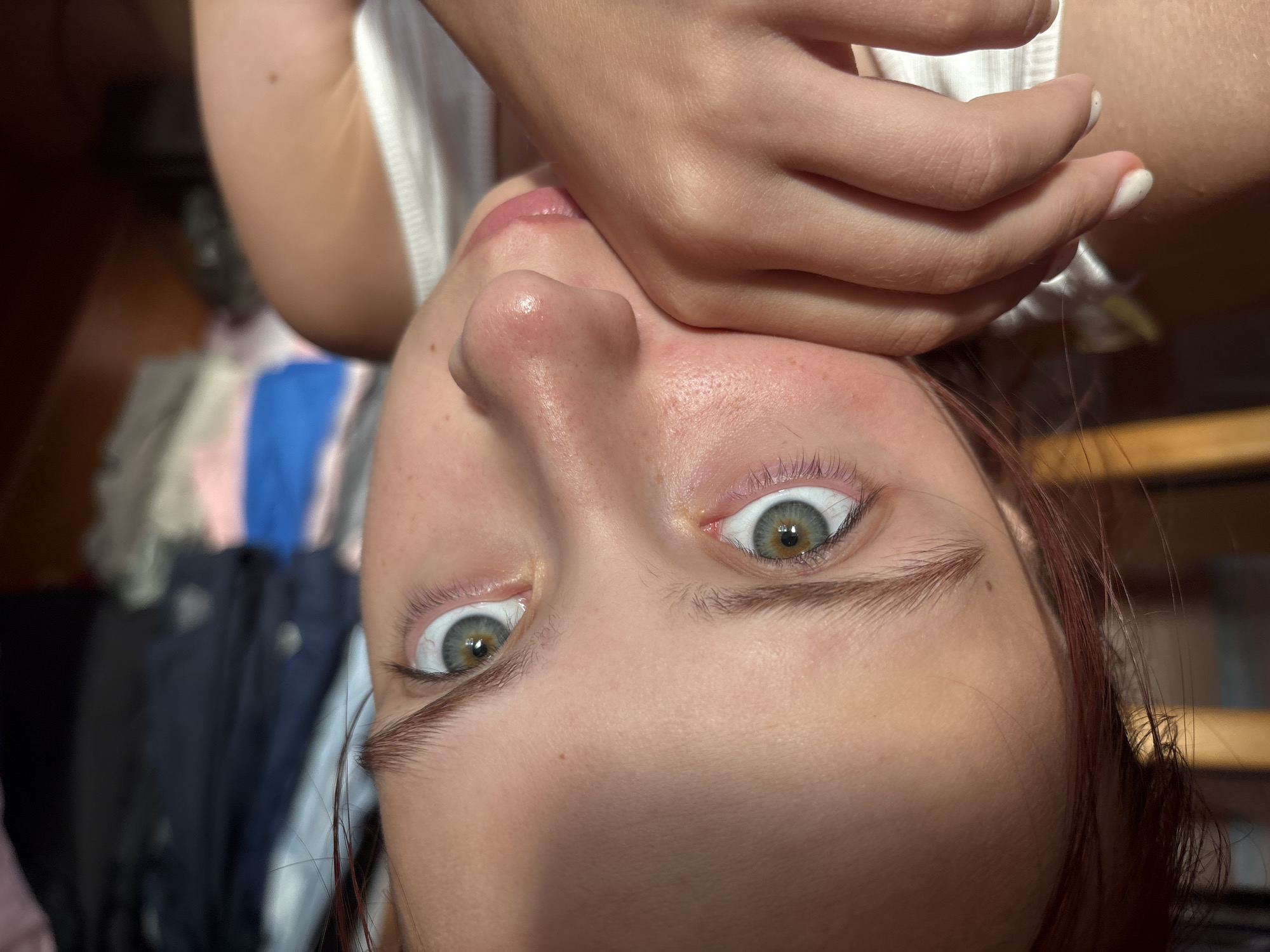 A close-up, indoor, medium-shot photograph shows a young female with fair skin and green eyes. Her right hand is resting on her chin, partially obscuring her mouth. Her head is tilted slightly to her left, and her gaze is directed at the camera. She has light brown hair that appears to be pulled back. She is wearing a white tank top. The background is blurred, with hints of clothing hanging on what appears to be a closet or rack. The lighting suggests natural light from the right side of the frame, casting shadows across her face. No specific location in the city of Прикамский, Russia can be determined from the visual information provided.
