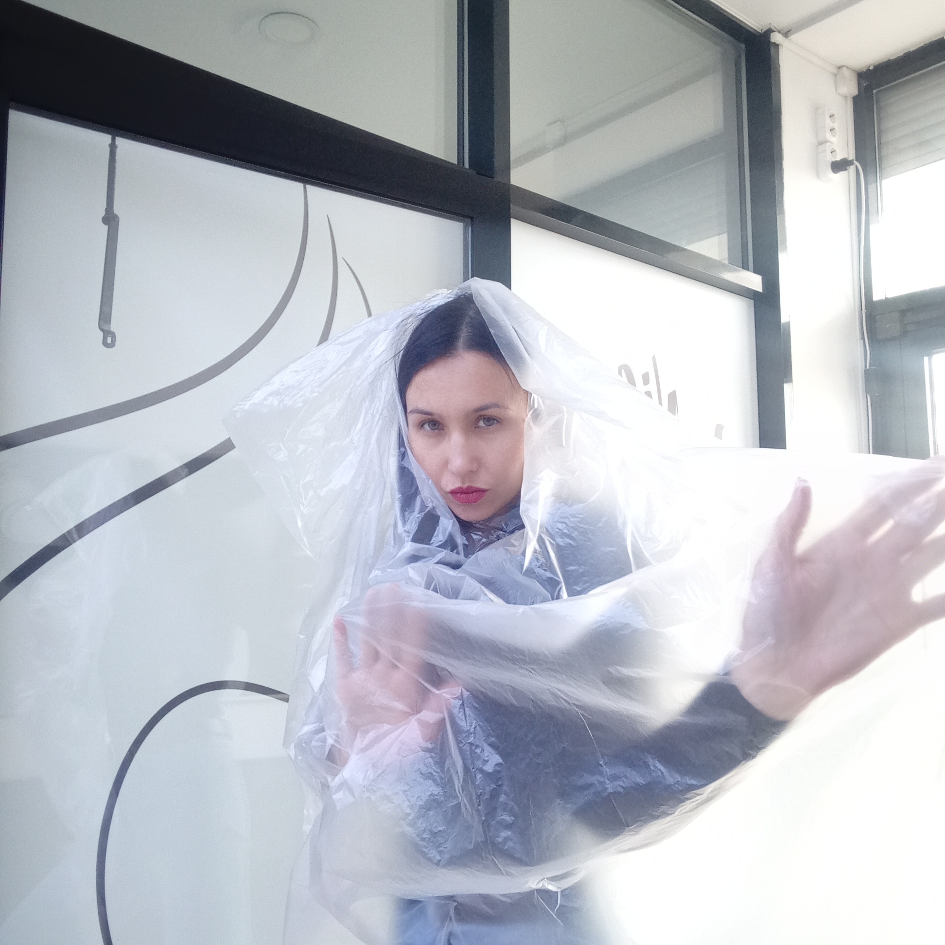 A portrait captures a young woman with dark hair parted in the middle and prominent red lipstick, looking directly at the camera. She is enveloped in clear, transparent plastic sheeting, which partially obscures her form and creates soft reflections and distortions. Her hands are visible through the plastic, extended outwards. She appears to be wearing a dark top or dress beneath the plastic.
The setting is an indoor space, likely a commercial or public building, featuring large windows or glass panels with dark frames. The background is brightly lit, suggesting daytime. On the light-colored surfaces behind her, there are decorative dark grey, stylized lines and a faint, partially visible, unreadable text or logo. To the upper left, a dark grey, slender mechanism, possibly a window stay, is attached to a panel. On the far right, a white wall includes a visible power outlet with a black plug inserted. The overall scene suggests a conceptual photoshoot, with the plastic serving as a key artistic element. The image was taken in Izhevsk, Russia.