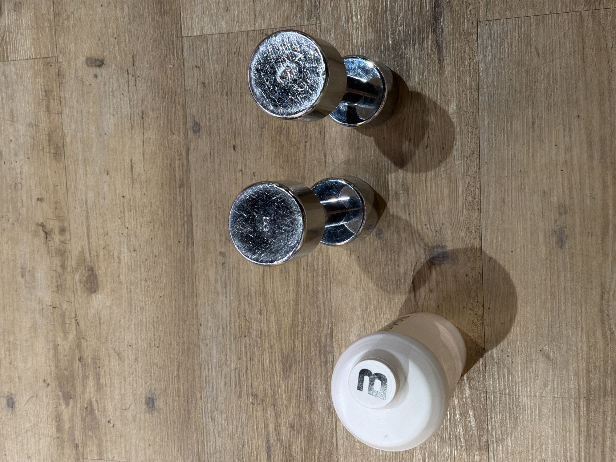 This image shows a white water bottle with a black "B" logo on its cap, and two silver dumbbells on a wooden floor. The water bottle is on the left side of the image, and the dumbbells are to the right. The wooden floor appears to be made of planks. There are no people visible in the image. The scene is indoors, and there is no indication of the specific location in Strasbourg, France.