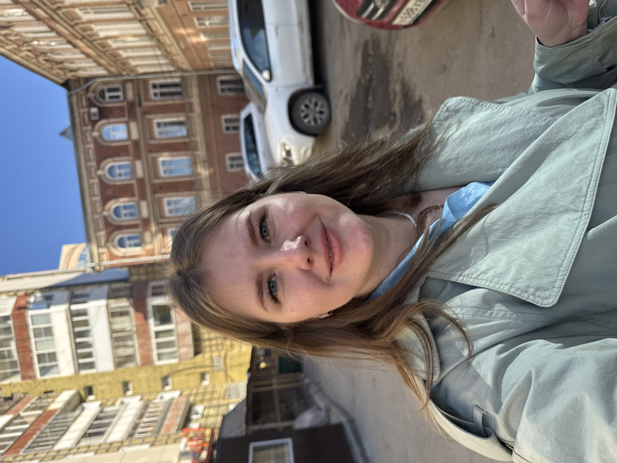 The image, rotated 90 degrees clockwise, captures a selfie of a young woman outdoors in an urban environment. The woman, likely in her 20s, has light brown hair, fair skin, and blue eyes, looking directly at the camera with a soft, pleasant smile. She wears a light blue top under a light green or grey long-sleeved jacket/trench coat and a pearl necklace.
In the background, several multi-story buildings with varied architectural styles and colors (reddish-brown, yellow, white) are visible under a clear blue sky. Two white cars are parked behind her, with a partial view of a red car's front bumper/fender in the upper right. Visible Cyrillic characters on the red car's license plate include "EX 885".
The scene takes place during a sunny day, likely late morning or early afternoon, indicating fair weather. The setting is a street or sidewalk in an older city, consistent with the provided context of Perm, Russia.