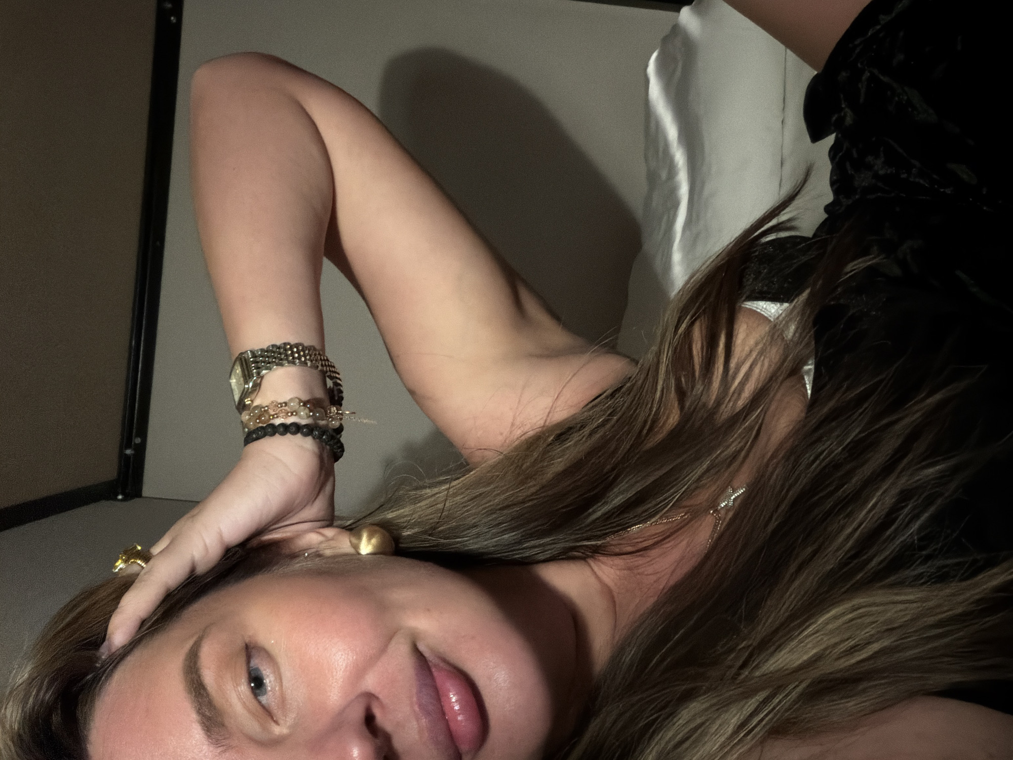 The image shows a woman lying on her side, with her head near the bottom of the frame. She has fair skin, brown hair, and a blue eye visible. Her lips are slightly parted, and she is wearing jewelry, including a watch and several bracelets on her wrist, and earrings. A gold ring is visible on her forehead. She appears to be wearing a black garment and there is a white object behind her. The background includes a dark cabinet and a neutral-colored wall. There is no indication of the location being Moscow, Russia.
