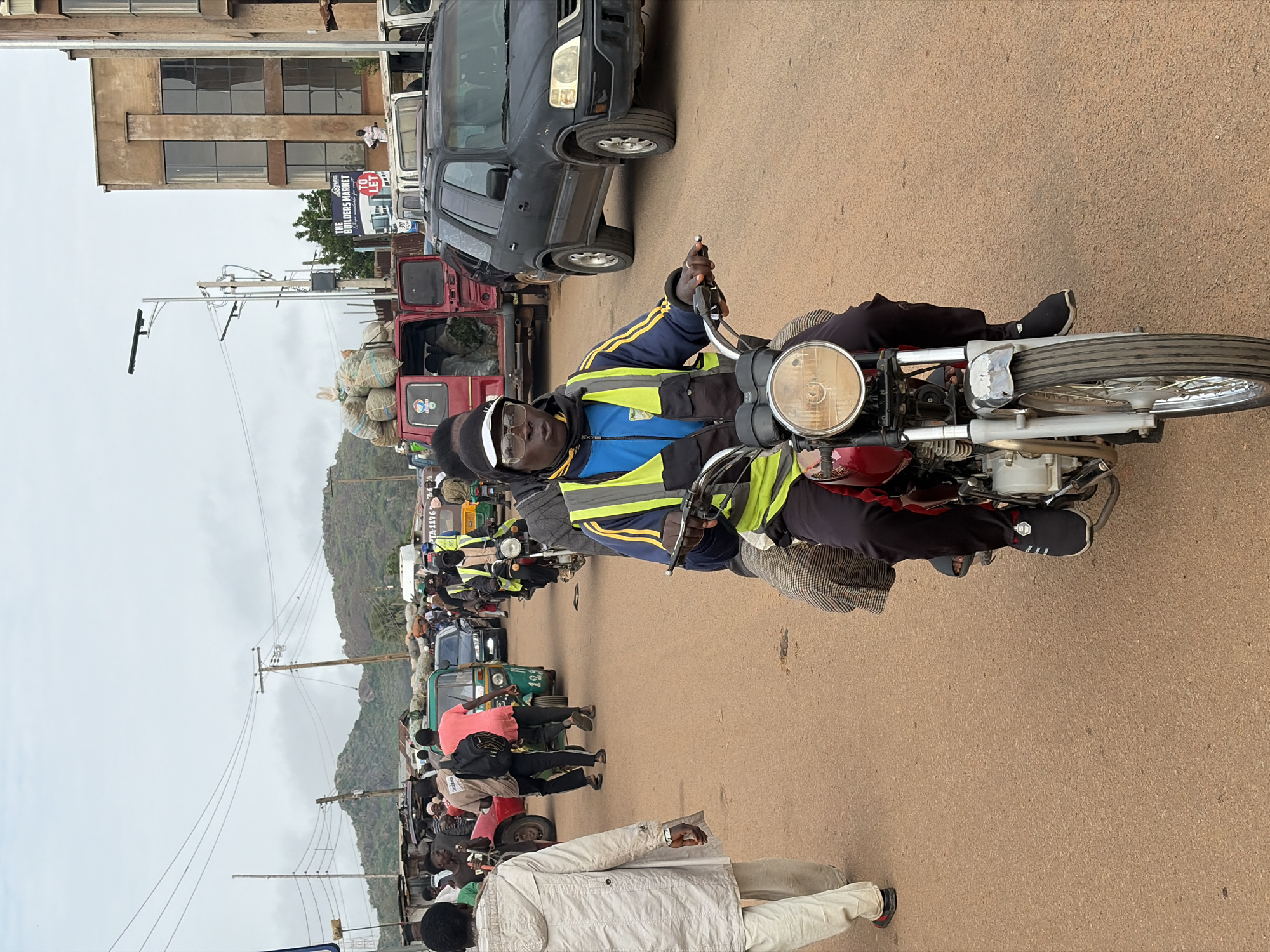 The image captures a busy street scene in Gura Top, Nigeria. The focal point is a man riding a motorcycle, wearing a blue jacket with yellow reflective stripes and a black cap. He is looking towards the camera. Behind him, traffic is congested with various vehicles including cars, a red van loaded with sacks of produce, and auto-rickshaws. Several people are seen walking and standing on the roadside, with market stalls and buildings lining the street. In the background, hilly terrain with sparse vegetation is visible under an overcast sky. The road surface is rough and dusty.