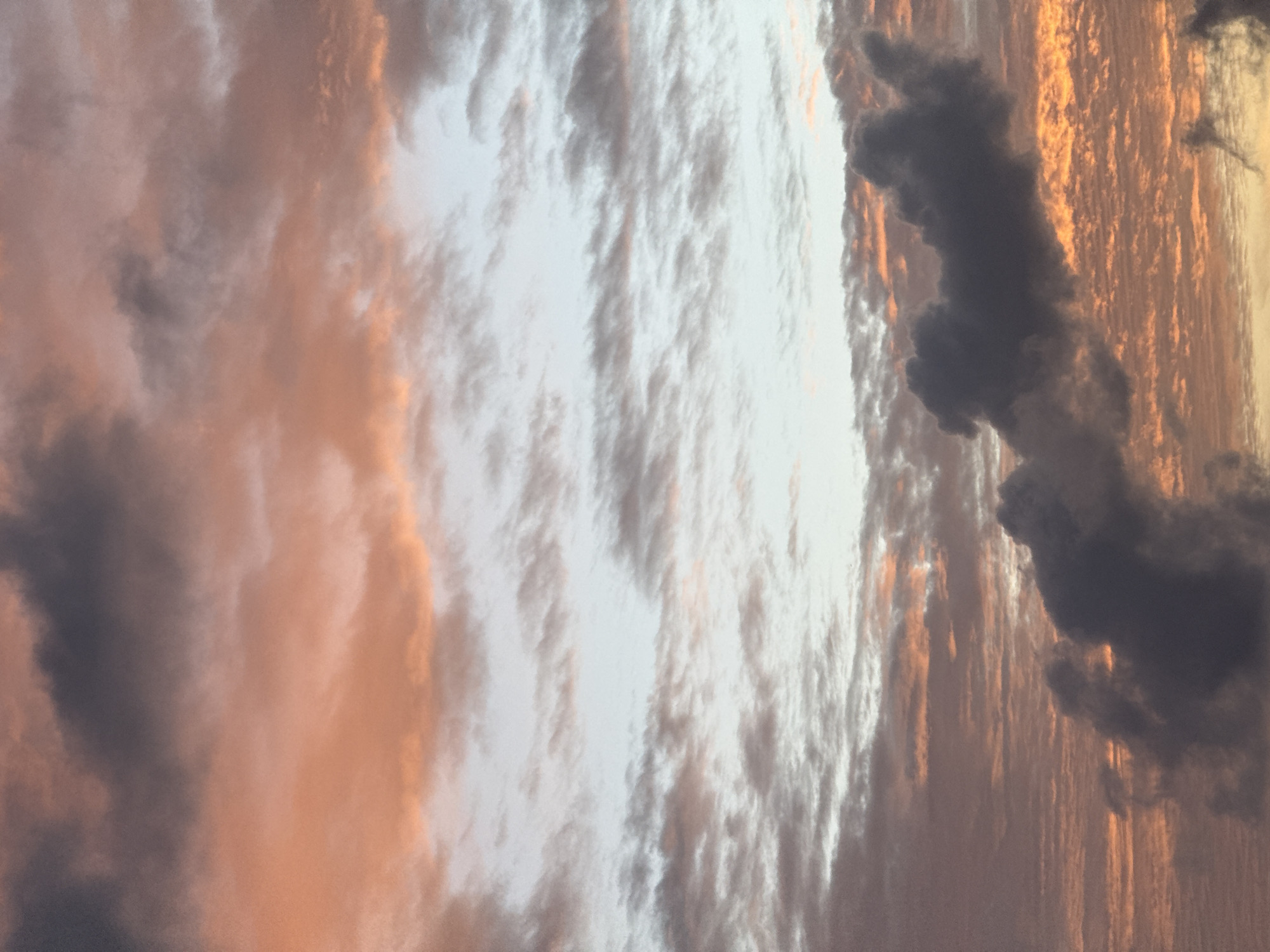 This is a photograph of a cloudy sky during what appears to be sunrise or sunset. The clouds are a mix of dark gray and light orange hues, suggesting the warm tones of the sun filtering through. The sky transitions from a pale blue at the top to deeper oranges and grays towards the bottom. There are no people, subjects, or objects visible in the image. The scene depicts a natural sky formation over the city of Karon, Thailand, characterized by diffuse cloud patterns and atmospheric lighting.