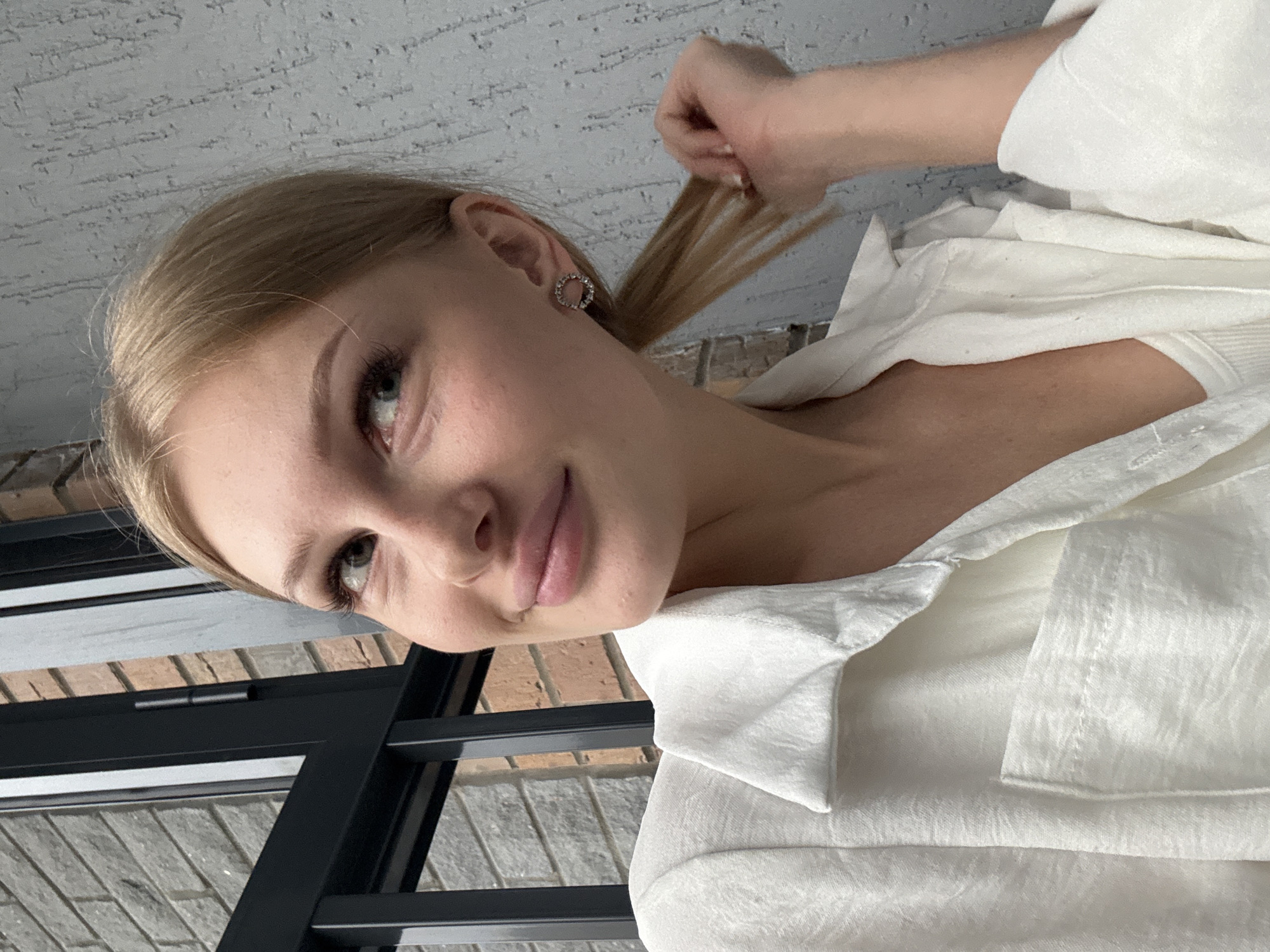 A young woman with blonde hair, wearing a white shirt and a white tank top, is captured from the chest up, looking to her left. She has a subtle smile and is touching her ponytail with her right hand. A circular earring with small stones adorns her left ear. The background consists of a textured grey wall and a brick wall visible through a black metal fence. The scene is indoors or on a covered balcony, with diffused lighting.