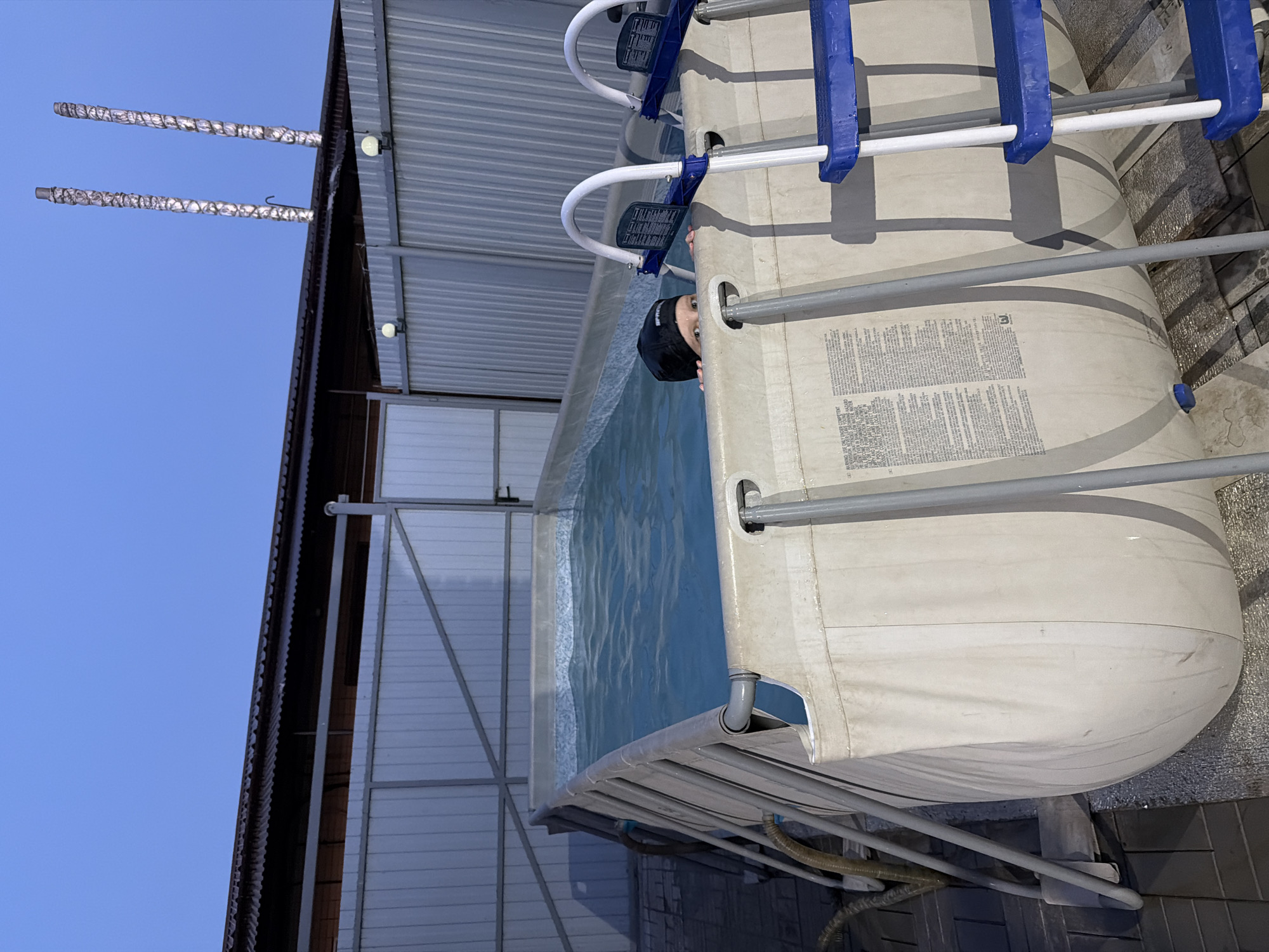An outdoor above-ground swimming pool is pictured with a person partially submerged in it. The pool is filled with blue water and has a white liner with text warnings printed on it. Two blue and white ladders are attached to the pool. The pool is situated outdoors, near a corrugated metal fence and roof structure. Two tall, insulated pipes are visible in the background, extending upwards against a clear blue sky. The ground around the pool appears to be paved with dark square tiles.