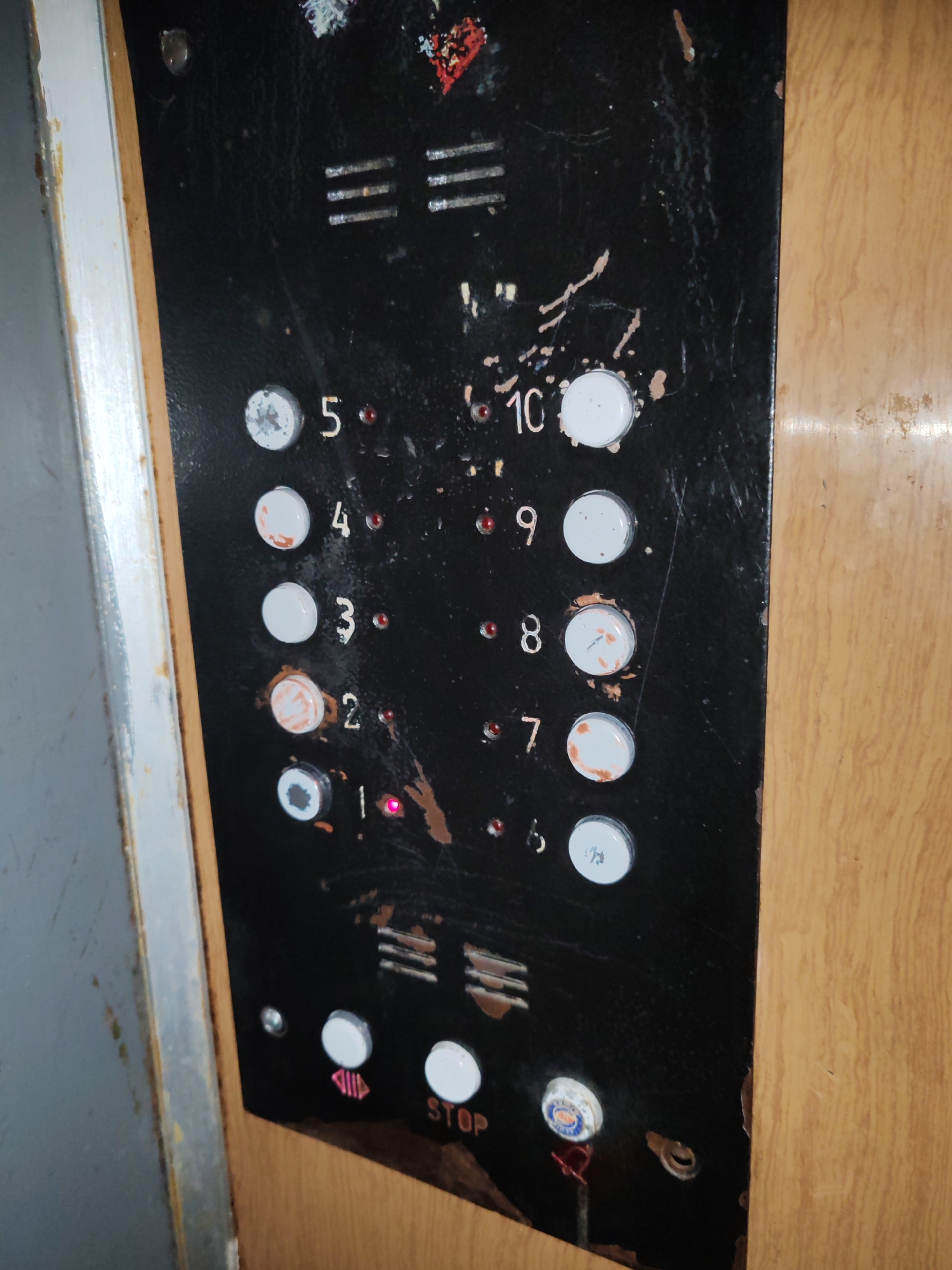 The image shows an old, worn elevator control panel. The panel is black with white numbering for the floors, ranging from 1 to 10. Several buttons are visible, some of which are lit. There are also two larger buttons at the bottom, one with an upward-pointing arrow and the other labeled "STOP". The panel is scratched and appears to have some rust or dirt on it. The surrounding environment suggests an interior setting, possibly a residential building or an older commercial establishment, with wooden paneling visible to the right. There is no information in the image to suggest a specific location in Poltava, Ukraine.