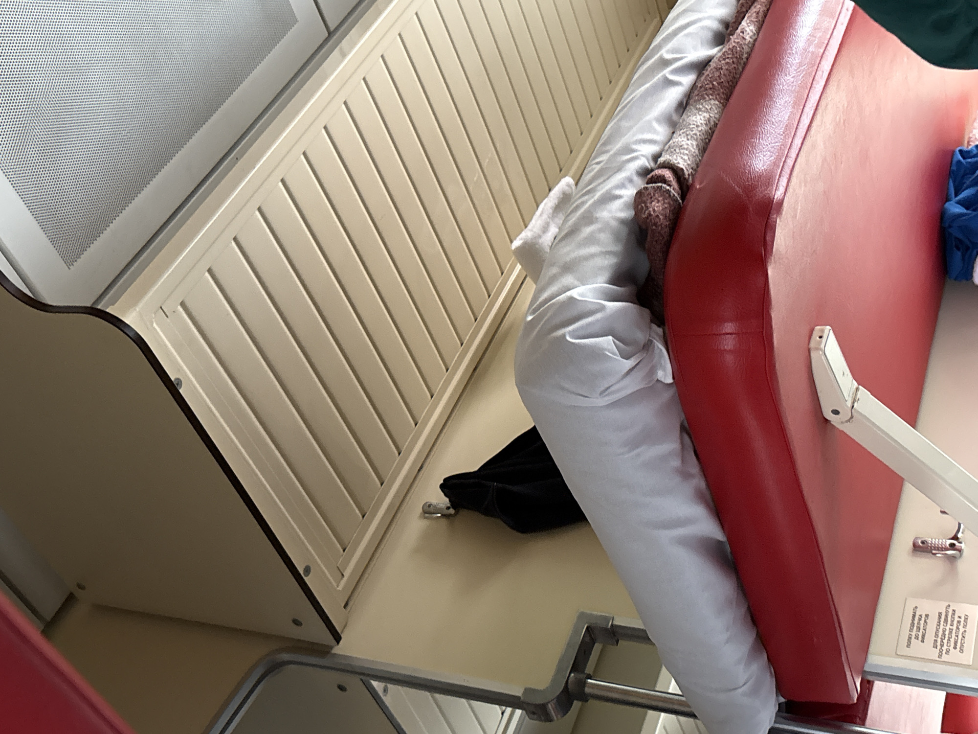 The image shows the interior of a train carriage, likely a sleeping compartment. The focus is on the upper bunk, which is made up with white bedding and a patterned blanket. A black jacket is hanging on a hook on the wall. The walls are painted a pale yellow, and the ceiling has horizontal slatted panels. A red upholstered seat is visible in the foreground. The overall impression is one of a functional and somewhat dated train interior. The image is taken from a low angle, looking up towards the bunk and ceiling.