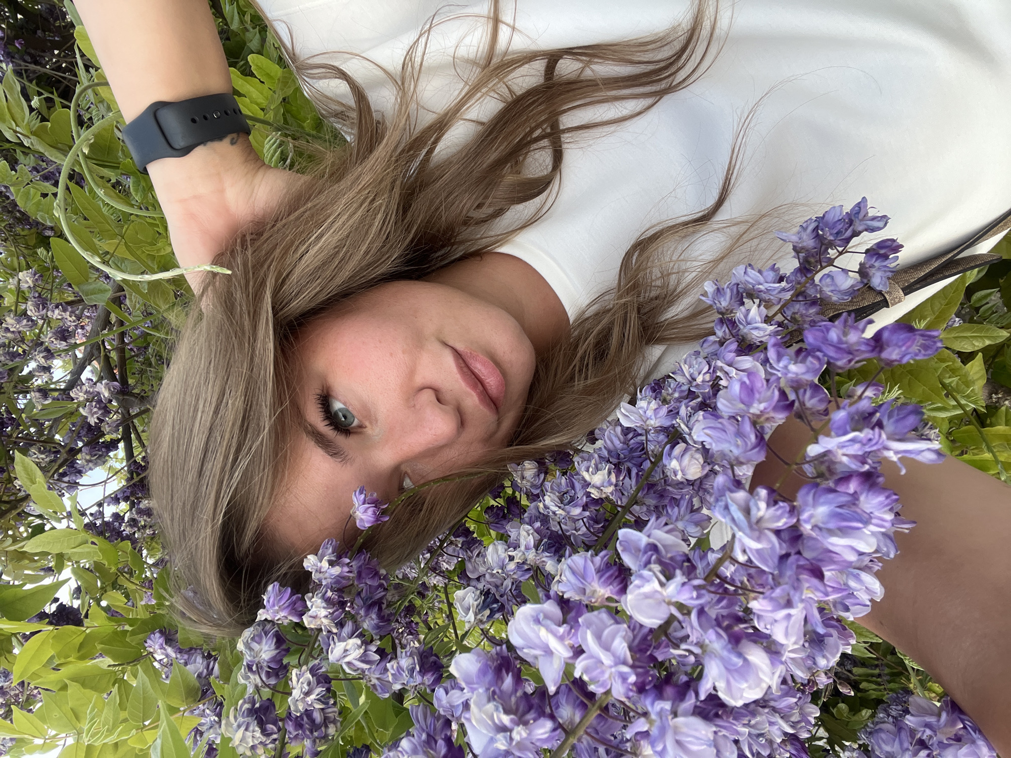 A woman is reclining amidst blooming wisteria flowers. She wears a white shirt. A black smart watch is visible on her left wrist. The location is likely a garden setting in Budva, Montenegro, given the presence of wisteria, a common ornamental plant in the region.
