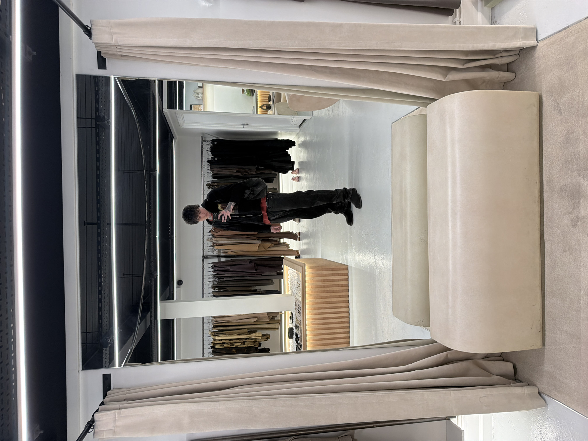 A person stands in what appears to be a fitting room within a store, possibly in London, United Kingdom. The person, likely male, is dressed in dark clothing, including a jacket and pants. They appear to be looking at themselves in the mirror, perhaps taking a photo with a phone. The room has a modern design with neutral tones. There is a clothing rack behind the person filled with various garments, primarily in shades of brown. A display cabinet with a light wood finish is also visible near the clothing rack. The fitting room itself is separated from the main store area by thick, light-colored curtains.