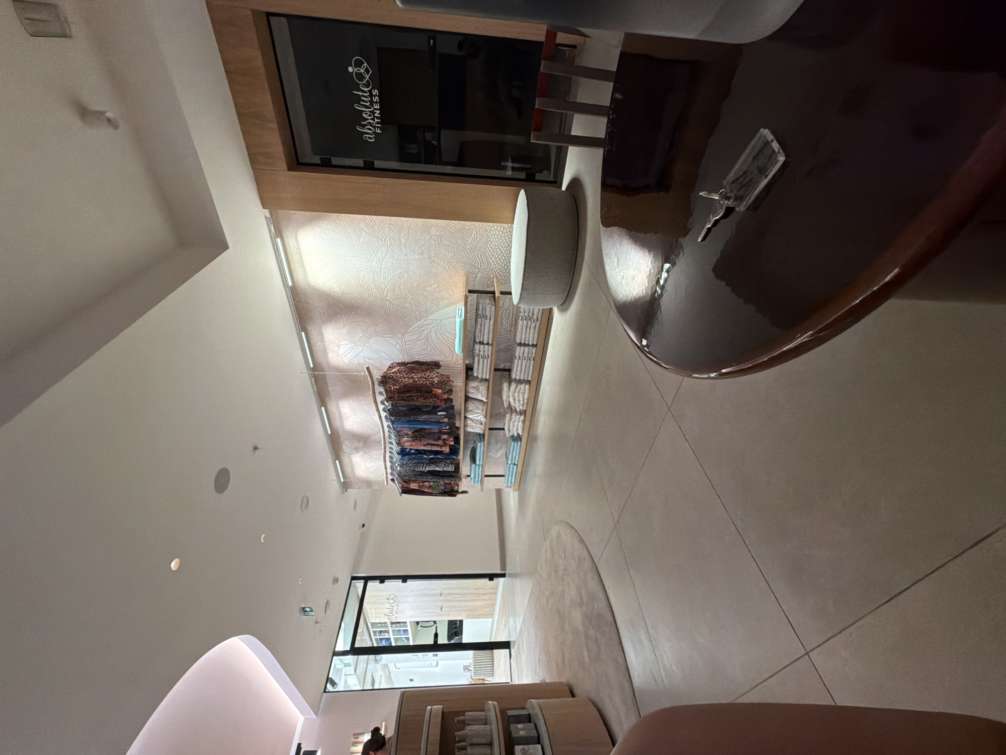 This is a wide-angle, eye-level shot of the interior of a retail space, possibly a boutique or studio, located in Boreti, Montenegro. The overall aesthetic is modern and clean, with light-colored tiled floors and beige walls.
In the foreground, a dark, highly reflective circular table dominates the lower right quadrant. On its surface, a silver key with a clear rectangular tag is visible.
The midground shows a rack of clothing with colorful, patterned garments hanging from it. Below the clothing, two shelves are stacked with folded items, appearing to be towels or linens, in light blue and white. To the left of the clothing rack, a round, light-grey ottoman-like seating object is positioned on the floor.
Further to the left, a curved, light-pink architectural element is visible, possibly part of the ceiling or a wall feature. Adjacent to this, a circular, beige rug is partially visible on the floor. A cylindrical shelving unit with multiple tiers holding various bottles and containers stands to the far left.
In the background, a doorway with a glass-paned door reveals a glimpse of another area, possibly a changing room or a back office, with wooden doors. To the right of this doorway, a large, dark glass door is present, displaying a white logo and text that reads "absolute FITNESS".
The lighting is bright and diffused, coming from ceiling lights and possibly accent lighting behind the clothing display, creating a well-lit and airy atmosphere. The scene is devoid of any people, suggesting the store is either closed or empty at the time the photograph was taken. The architectural elements, particularly the curved pink structure and the contrast between the modern signage and the more natural textures of the shelving and seating, suggest a contemporary design.