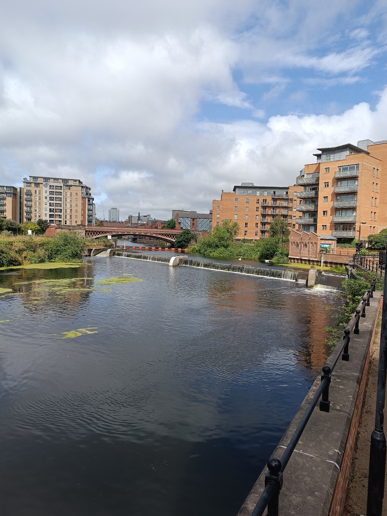 The image depicts a view of the River Aire in Leeds, United Kingdom, near the Holbeck area. In the foreground, the dark water of the river is visible, with some patches of green algae on the surface. A concrete embankment with a black metal railing runs along the right side of the river.
In the middle ground, a weir spans the width of the river, with water cascading over it. Beyond the weir, a red steel bridge with arched supports crosses the river, connecting the banks.
The background features modern residential buildings constructed from brick and glass, characteristic of urban development along the waterfront. These buildings are multi-story and appear to be apartments or condominiums. Greenery, including trees and bushes, is visible along the riverbanks between the water and the buildings. The sky is partly cloudy, with a mix of white clouds and patches of blue sky.
